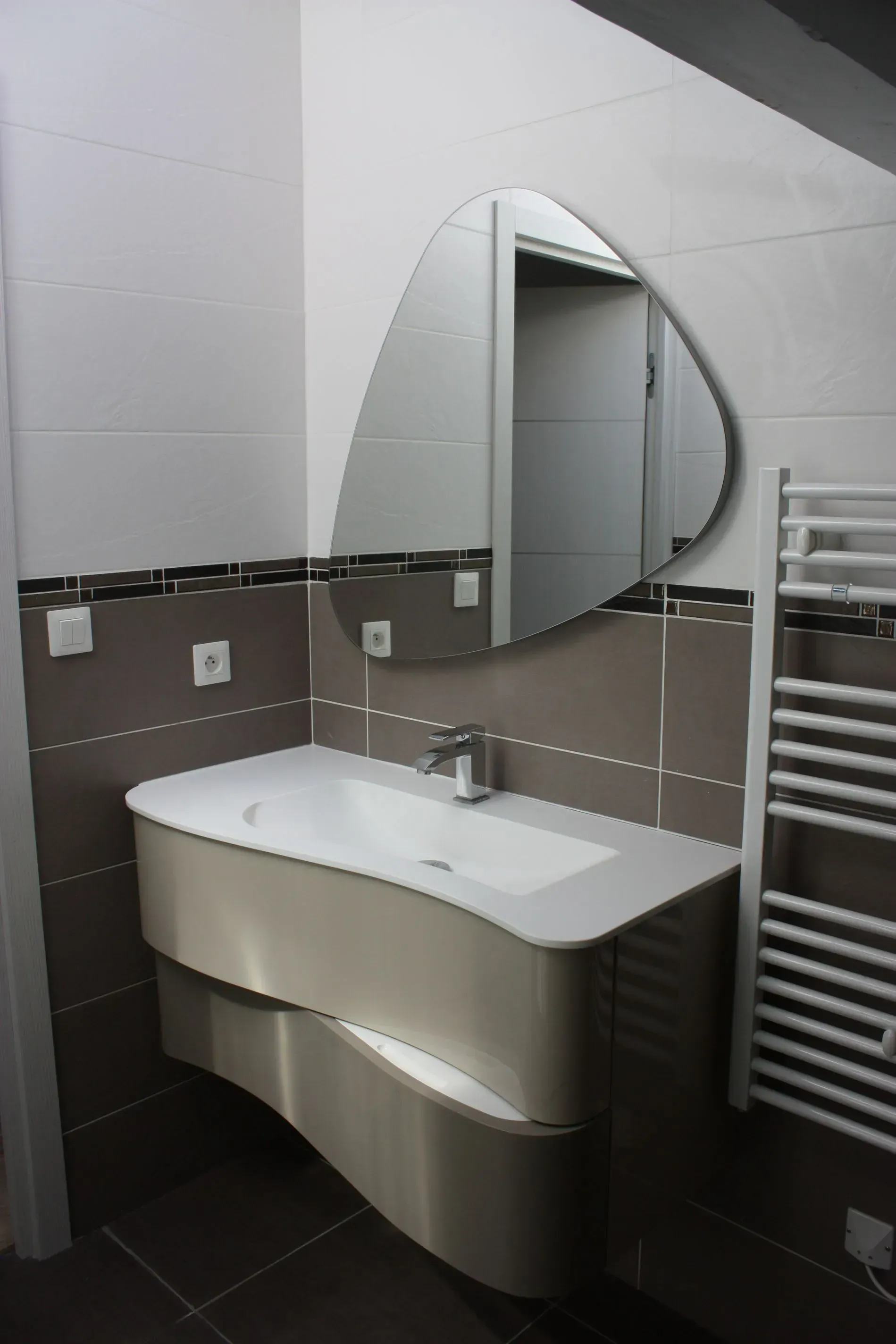 Un meuble lsous avabo moderne avec un meuble beige incurvé, un lavabo blanc et un miroir de forme irrégulière.