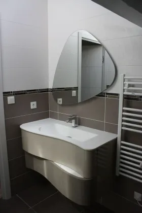 Un meuble lavabo moderne avec un meuble beige incurvé, un lavabo blanc et un miroir unique de forme irrégulière au-dessus.