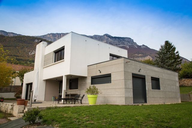 Une maison moderne de deux étages, blanche et en pierre, avec des volets sombres.