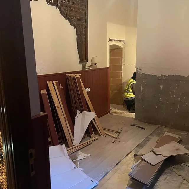 Un hombre con un chaleco amarillo está arrodillado en una habitación con mucha madera en el suelo.