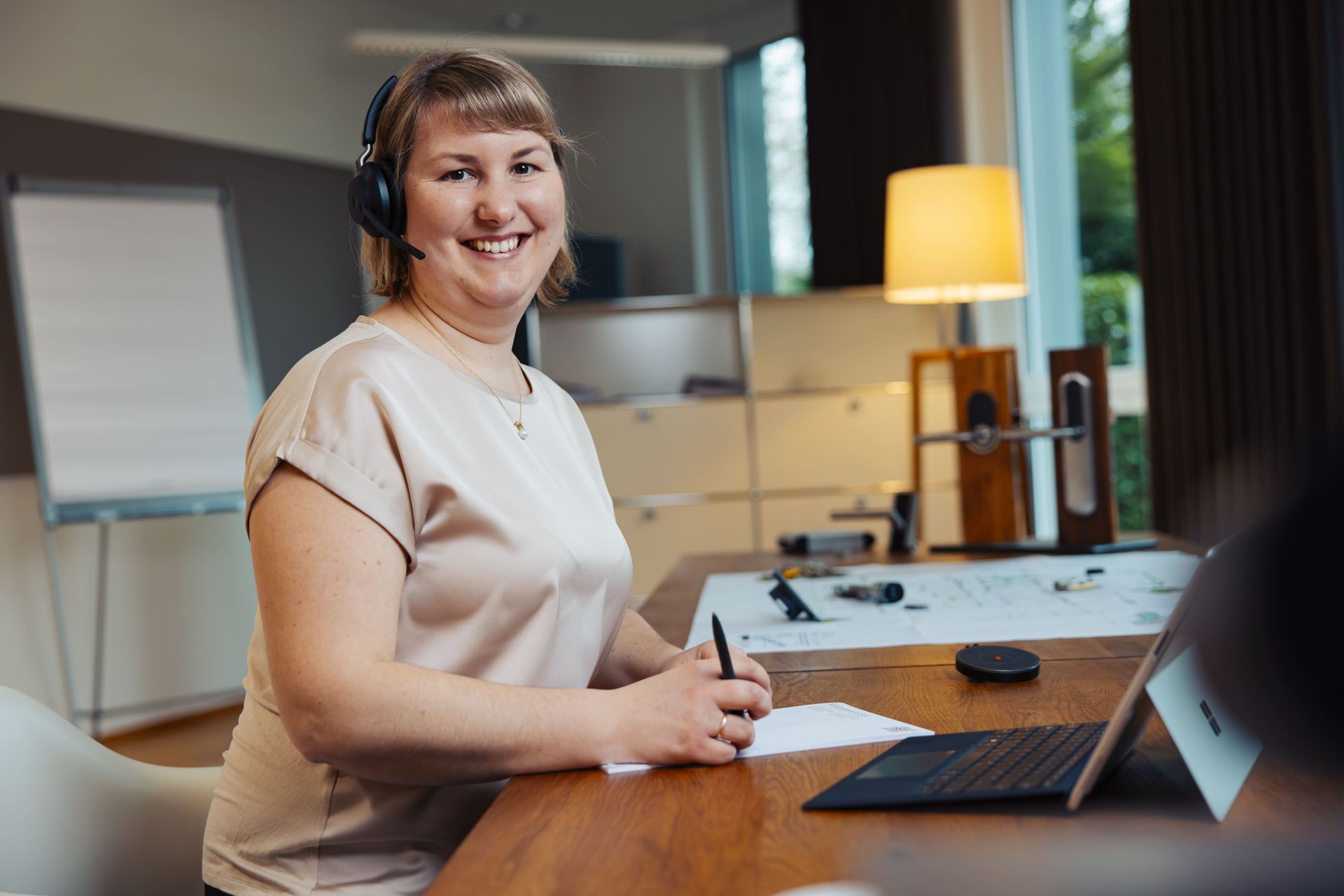Frau in schwarzem Blazer und Headset lächelt, während sie an einem Schreibtisch in einem gut beleuchteten Büro arbeitet.