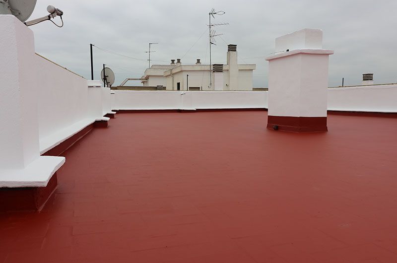 Terraza en la azotea con suelo rojo, paredes blancas y edificios a lo lejos bajo un cielo nublado