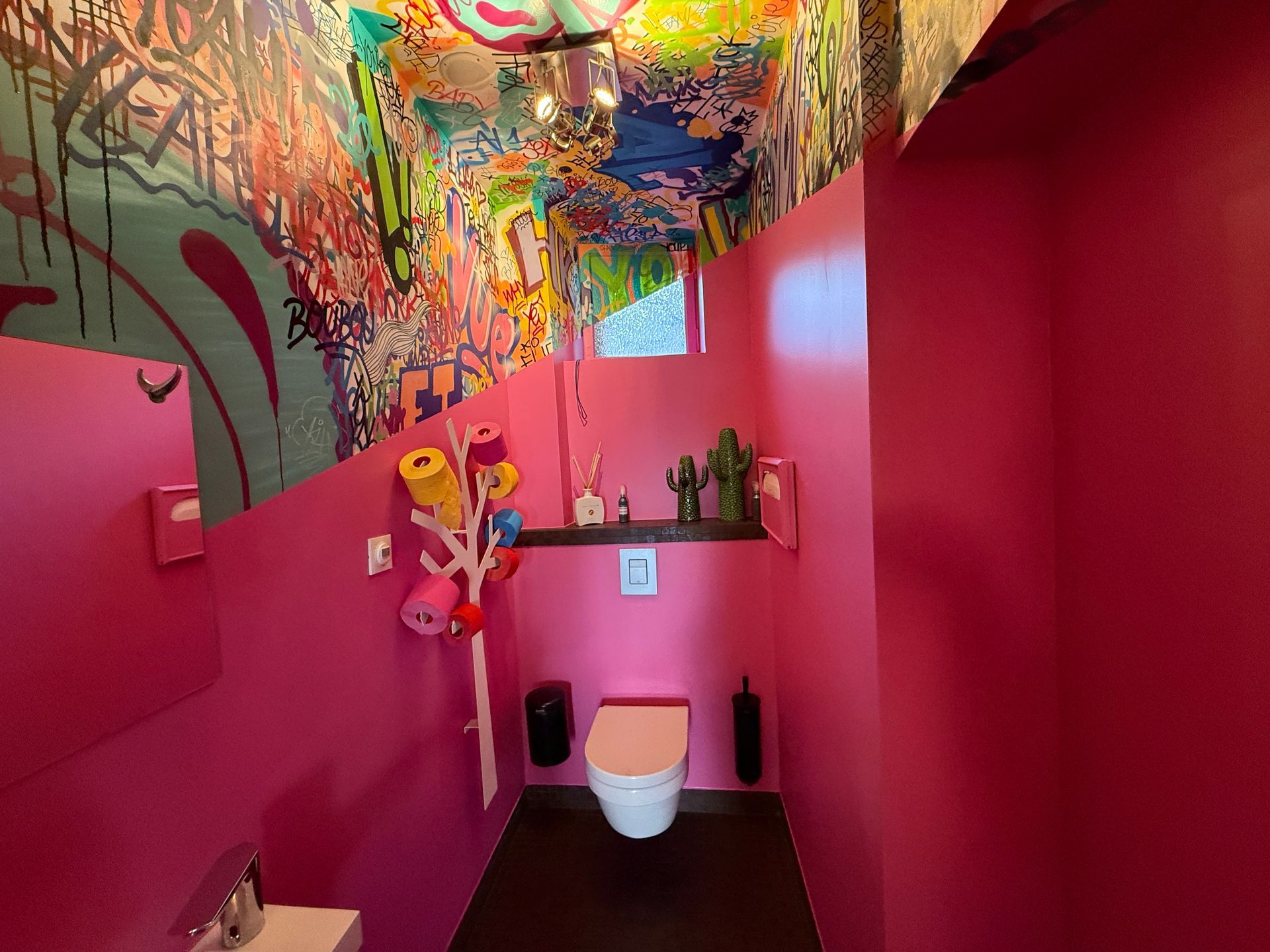 Intérieur des toilettes avec peinture rose et, à mi hauteur des murs et au plafond, des graffitis colorés