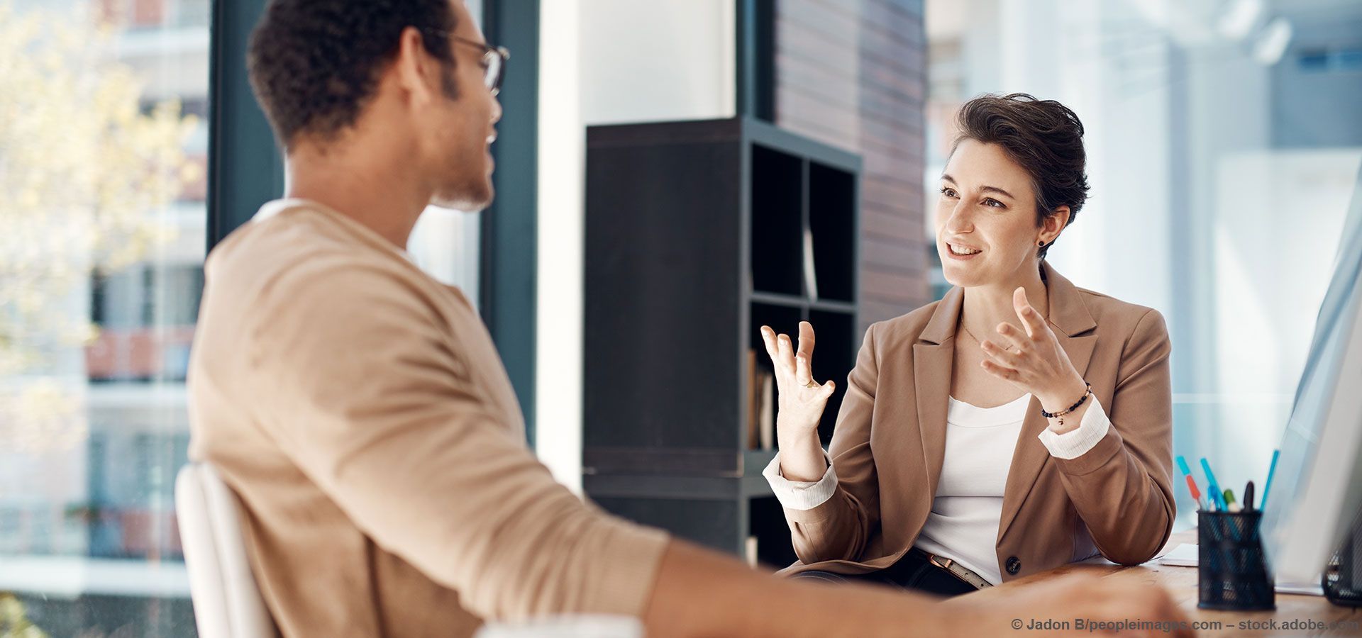Mann und Frau in einem Büro, die Frau gestikuliert und spricht mit dem Mann am anderen Ende des Schreibtischs.