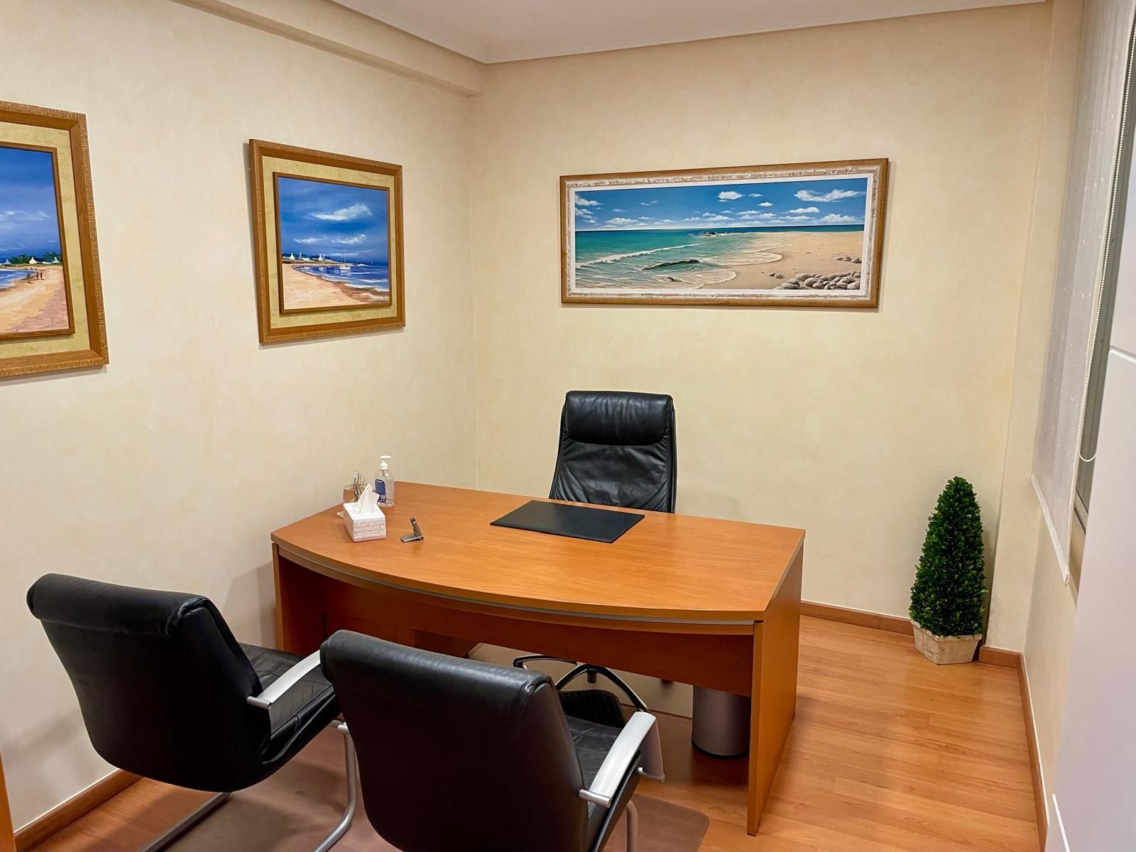 Pequeña oficina con escritorio de madera, sillas negras, cuadros en la pared y una planta en maceta en el suelo.