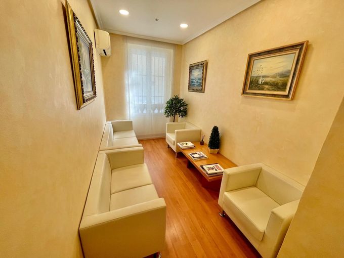 Pequeña sala de estar con sofás beige, suelo de madera, cuadros enmarcados y una ventana con cortinas transparentes.