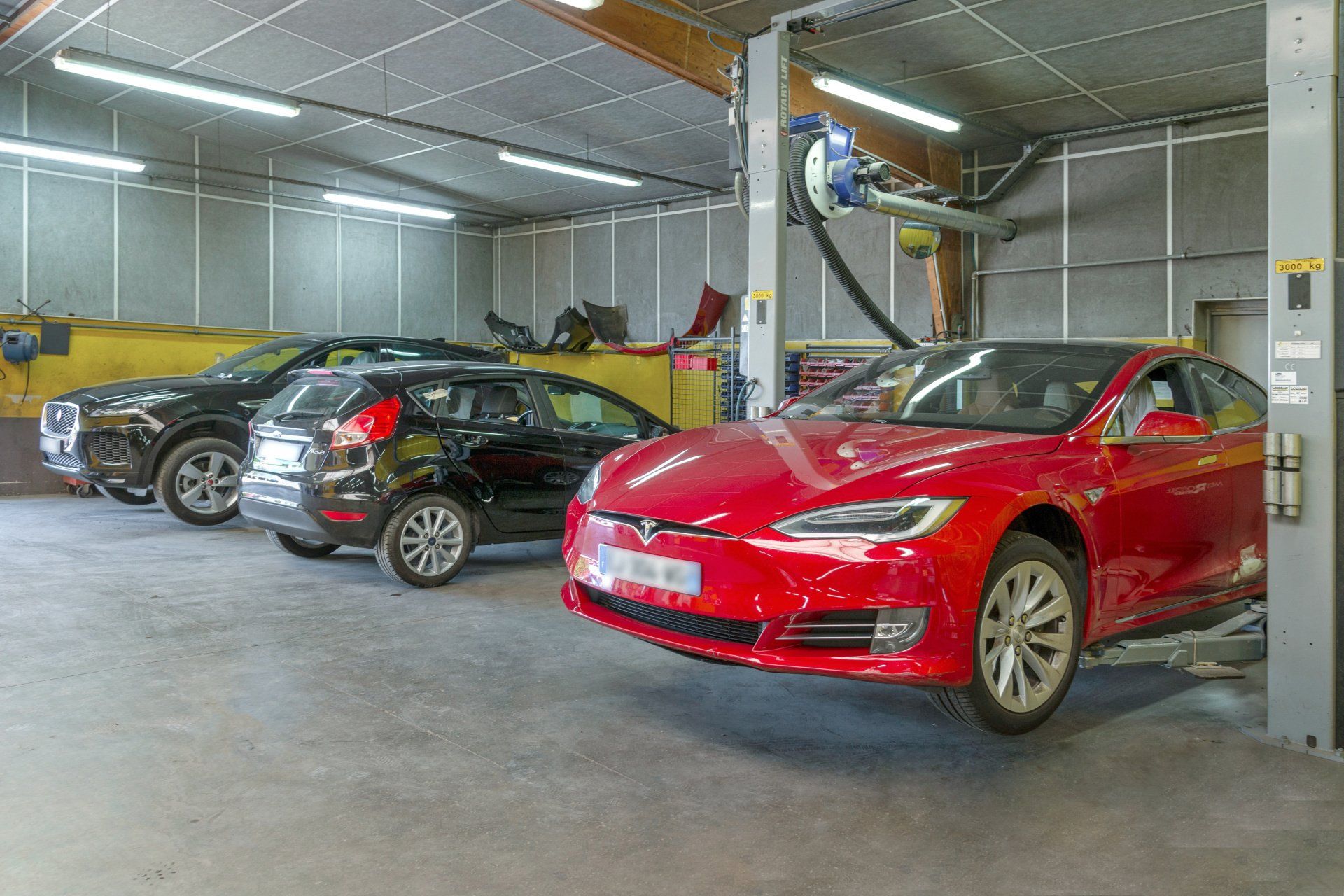 Vue de l'atelier de la Carrosserie Colas de l'intérieur, présentant trois voitures dont deux surélevées, en attente d'intervention.