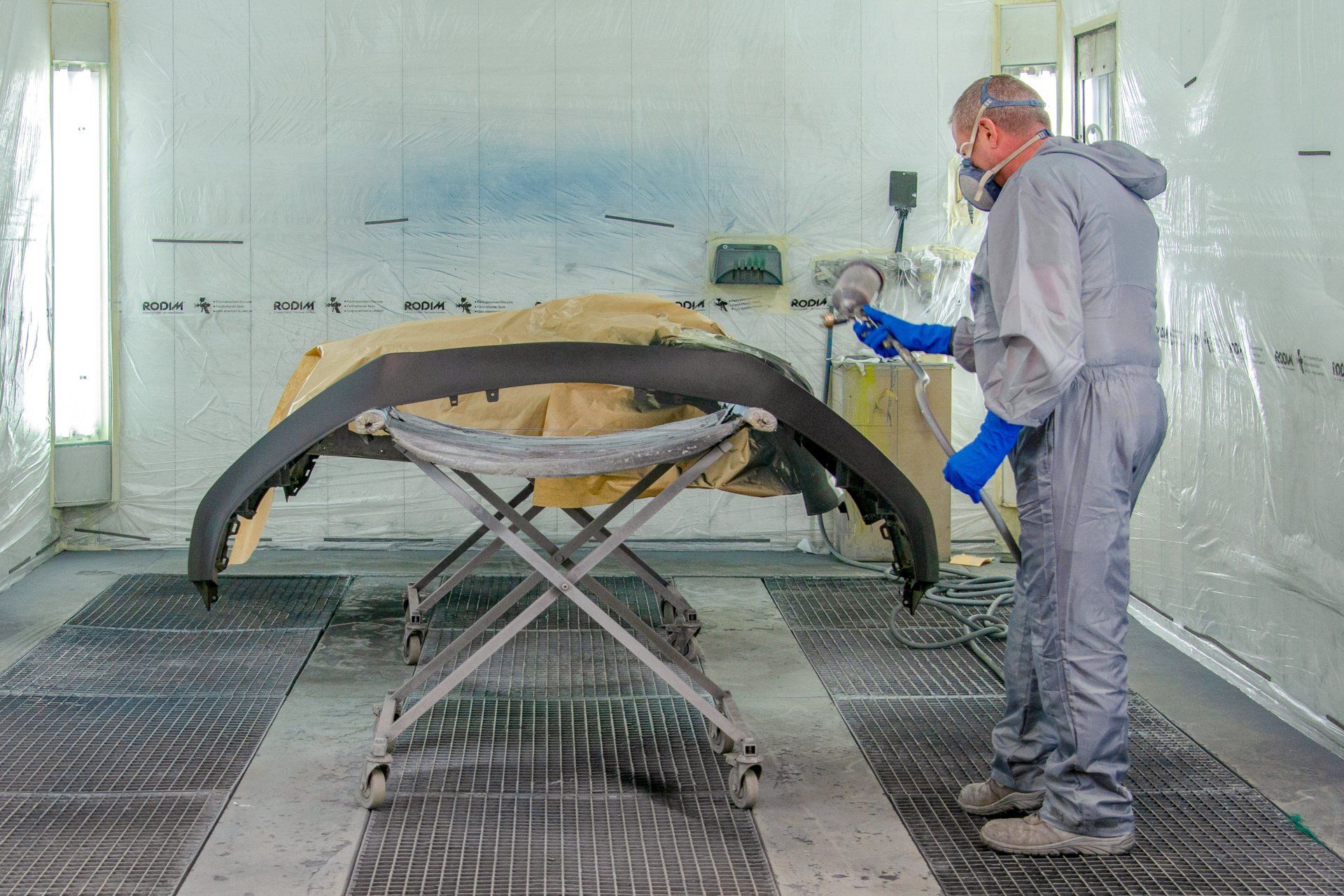 Pare-chocs en pleine opération de peinture par un technicien en combinaison grise placé à droite dans l'espace protégé de l'atelier.