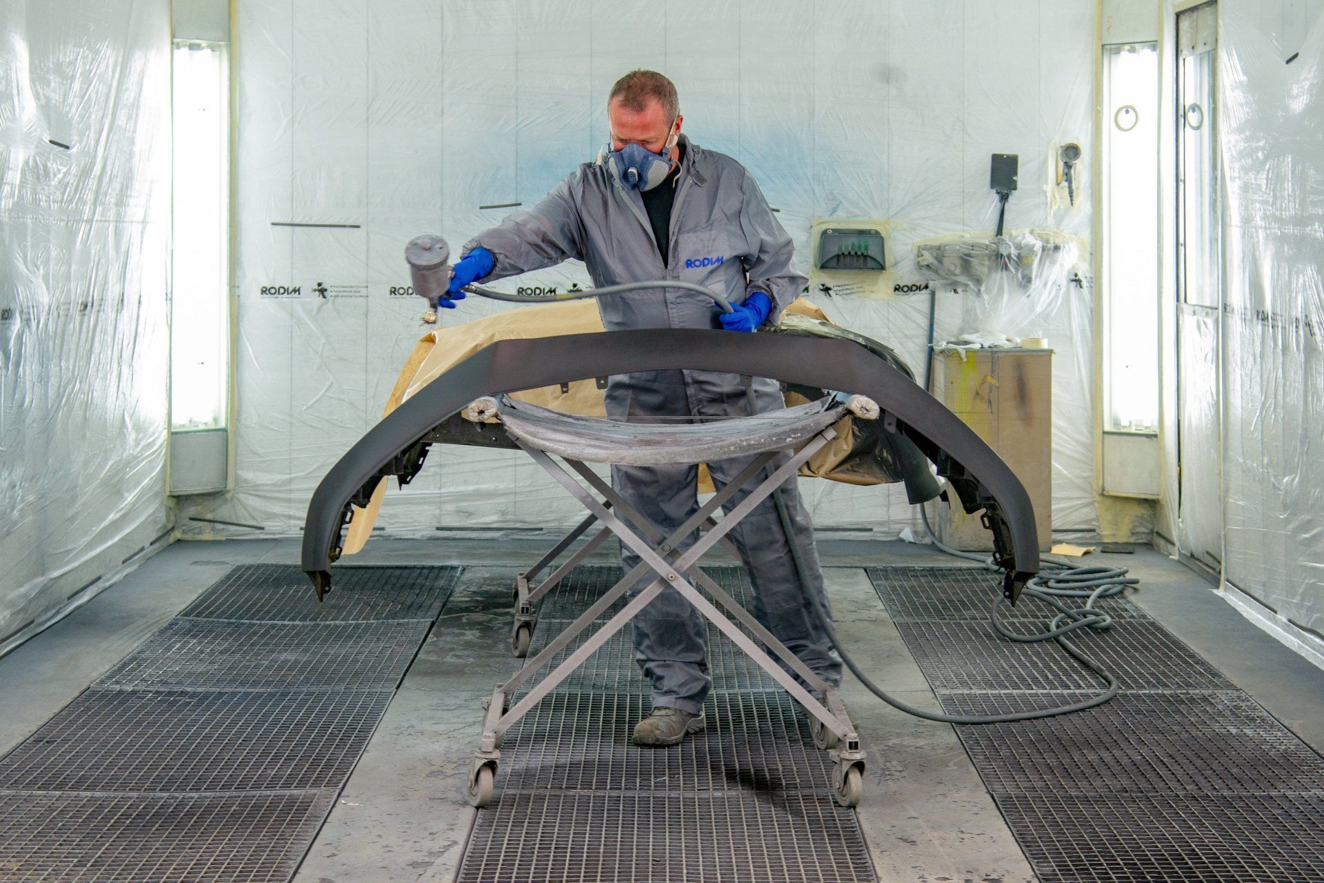 Vue sur l'espace protégé de l'atelier où un pare-chocs se fait repeindre par un technicien en combinaison.