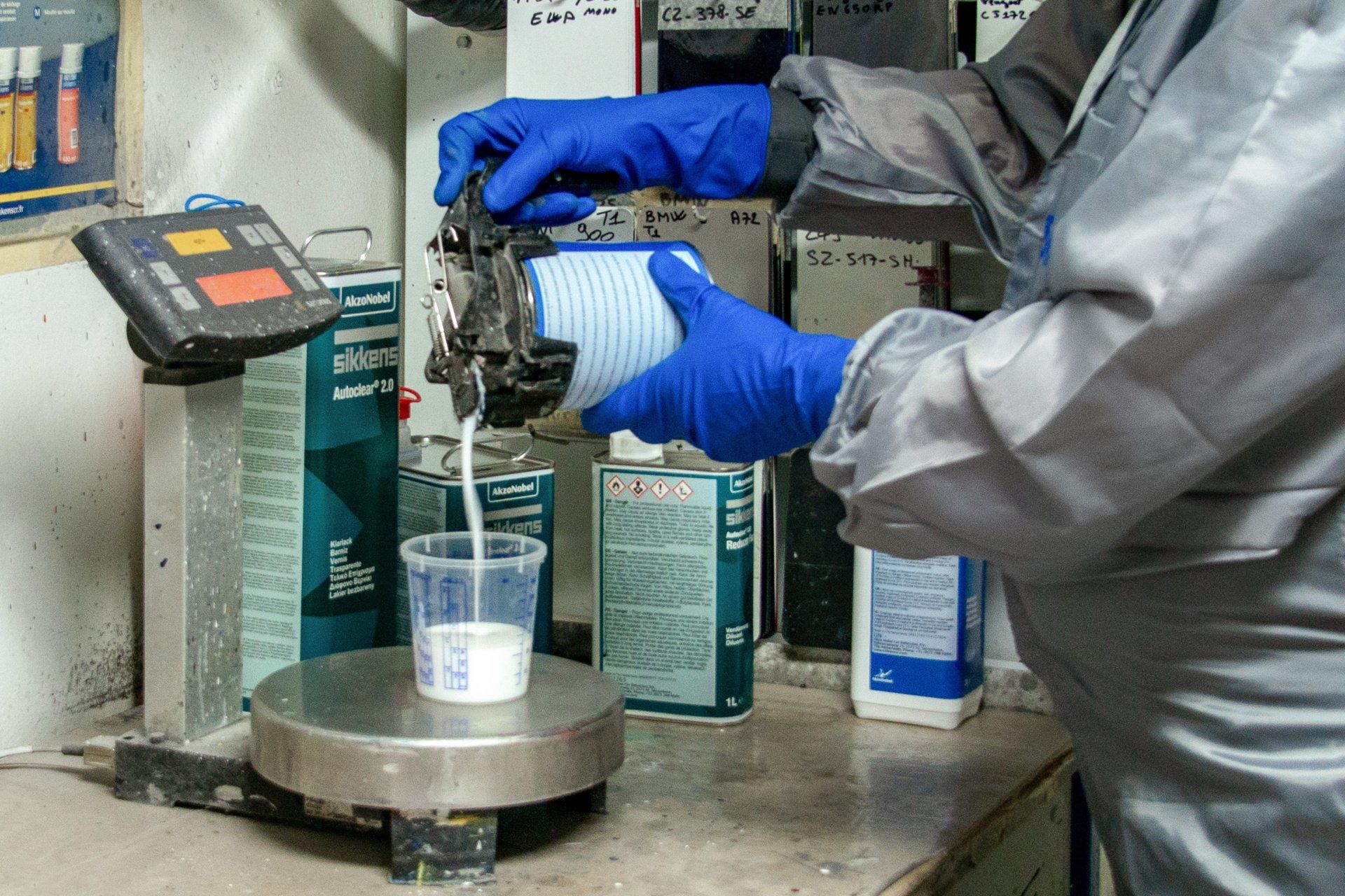 Zoom sur un technicien en combinaison grise qui verse un liquide blanc dans un verre doseur sur une balance.