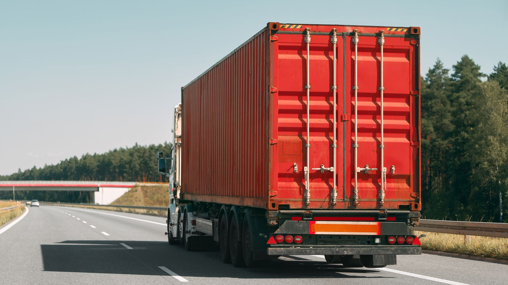 Un semi-remorque rouge transportant un conteneur de marchandises circule sur l'autoroute, avec des arbres et un pont en arrière-plan.