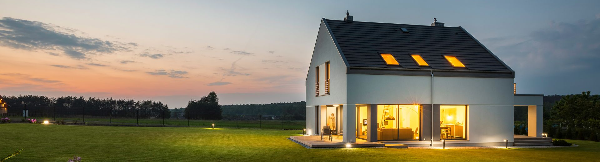 Maison blanche moderne au crépuscule, avec fenêtres illuminées et pelouse.
