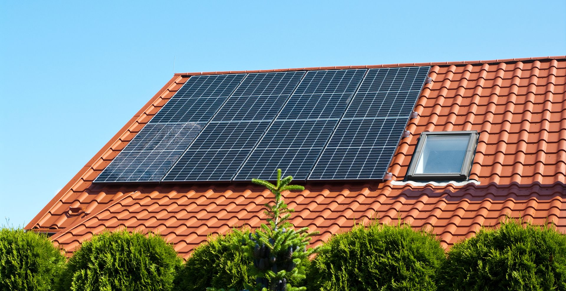Des panneaux solaires sur un toit de tuiles rouges percé d'un puits de lumière, se détachant sur un ciel bleu, au-dessus de buissons verdoyants.