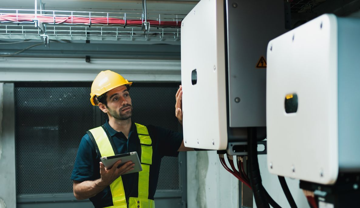 Un électricien portant un casque de chantier inspecte un onduleur solaire à l'aide d'une tablette dans un environnement industriel.