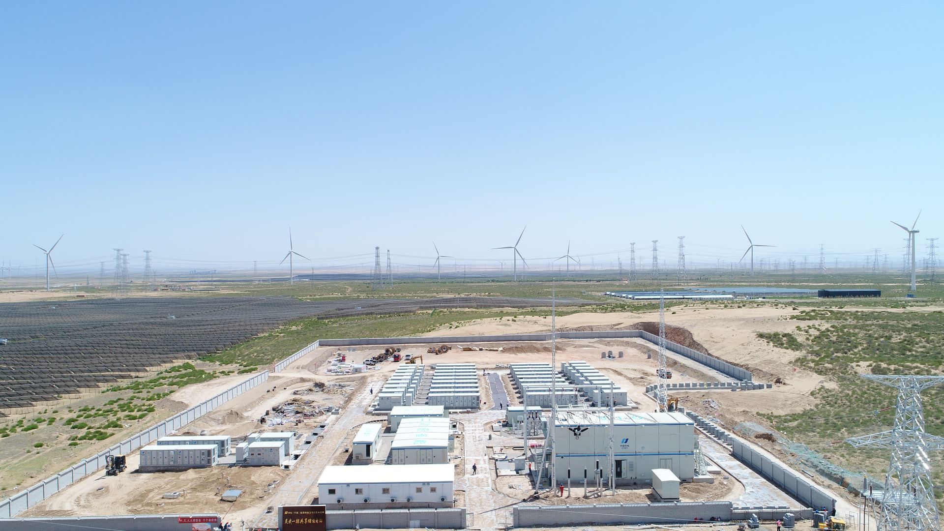 Vue aérienne d'un poste de transformation électrique avec des éoliennes en arrière-plan, sous un ciel bleu.