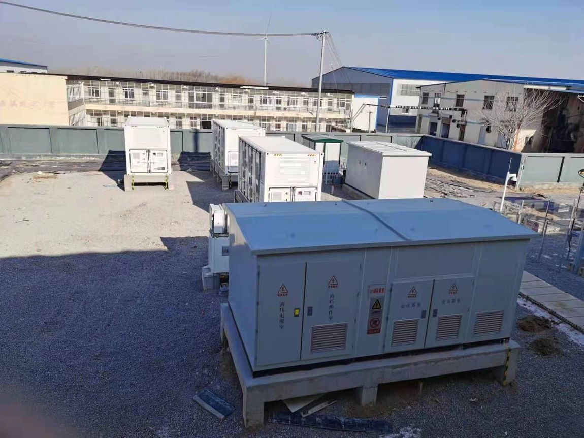Équipements électriques extérieurs, boîtiers et conteneurs gris sur un terrain gravillonné près de bâtiments, sous un ciel dégagé.