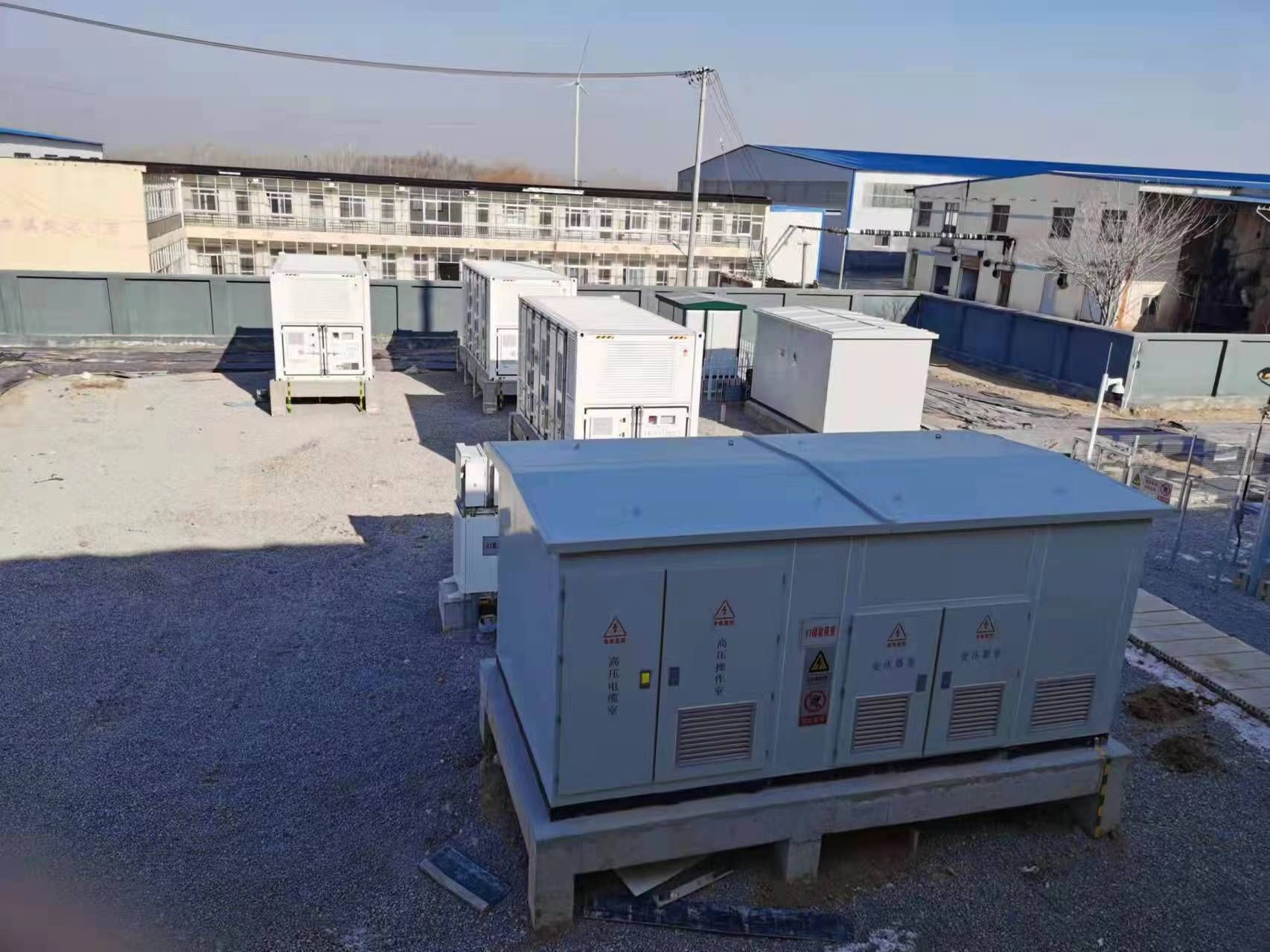 Équipements électriques extérieurs, boîtiers et conteneurs gris sur un terrain gravillonné près de bâtiments, sous un ciel dégagé.