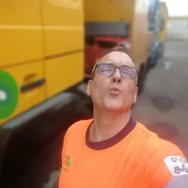 Un hombre que lleva gafas y una camiseta naranja se toma una selfie delante de un camión amarillo.