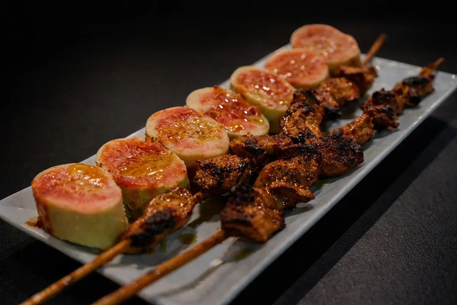 Comida a la parrilla en brocheta con higos, sobre un plato rectangular blanco.
