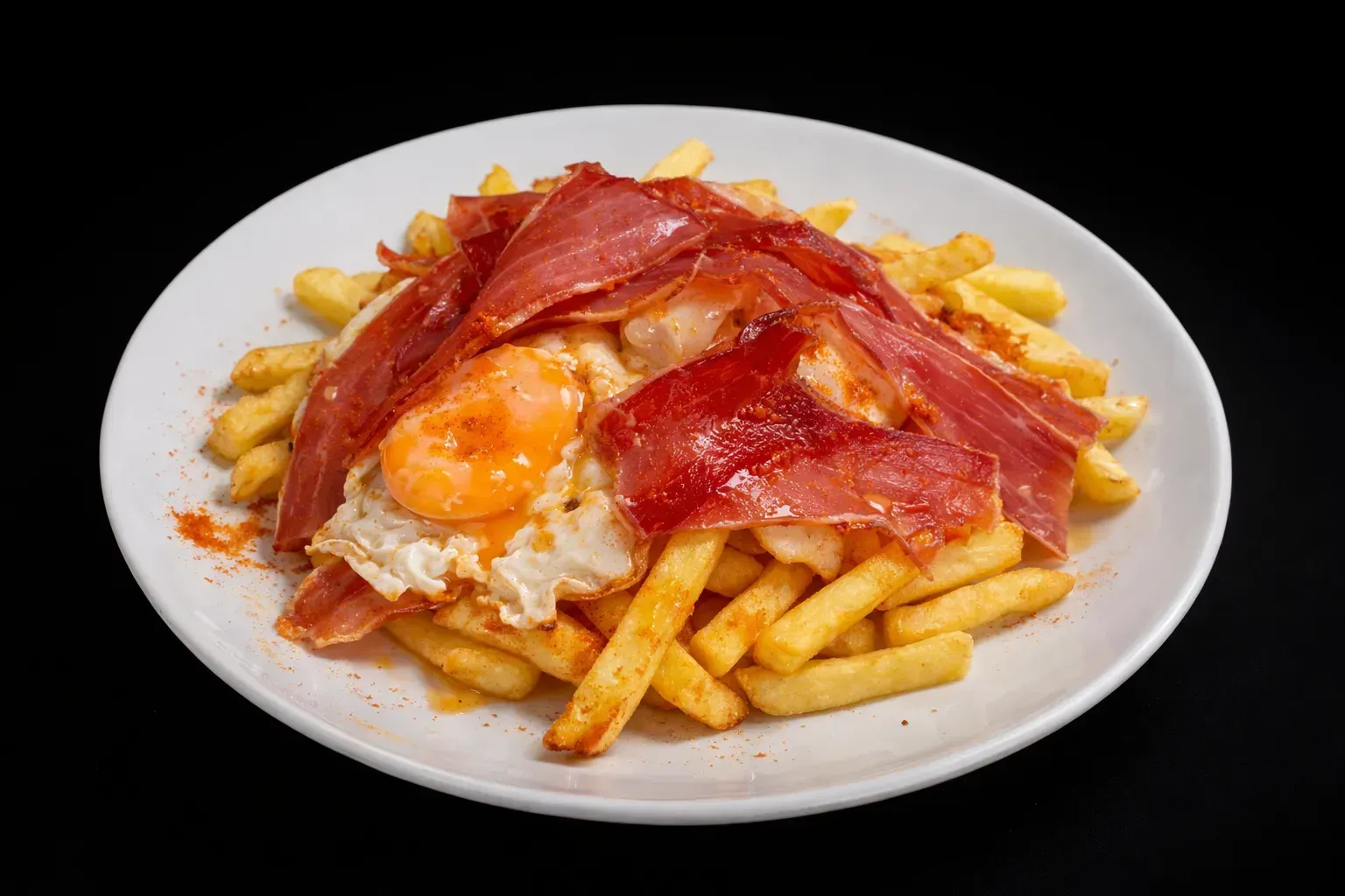 Papas fritas cubiertas con un huevo frito y jamón serrano en rodajas finas en un plato blanco.
