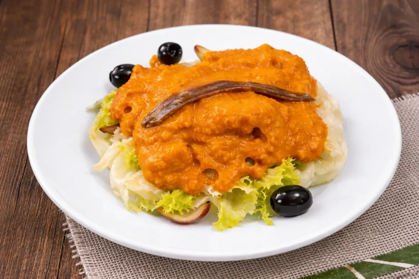 Plato blanco con pan cubierto con salsa roja, una anchoa, aceitunas y lechuga sobre una mesa de madera.
