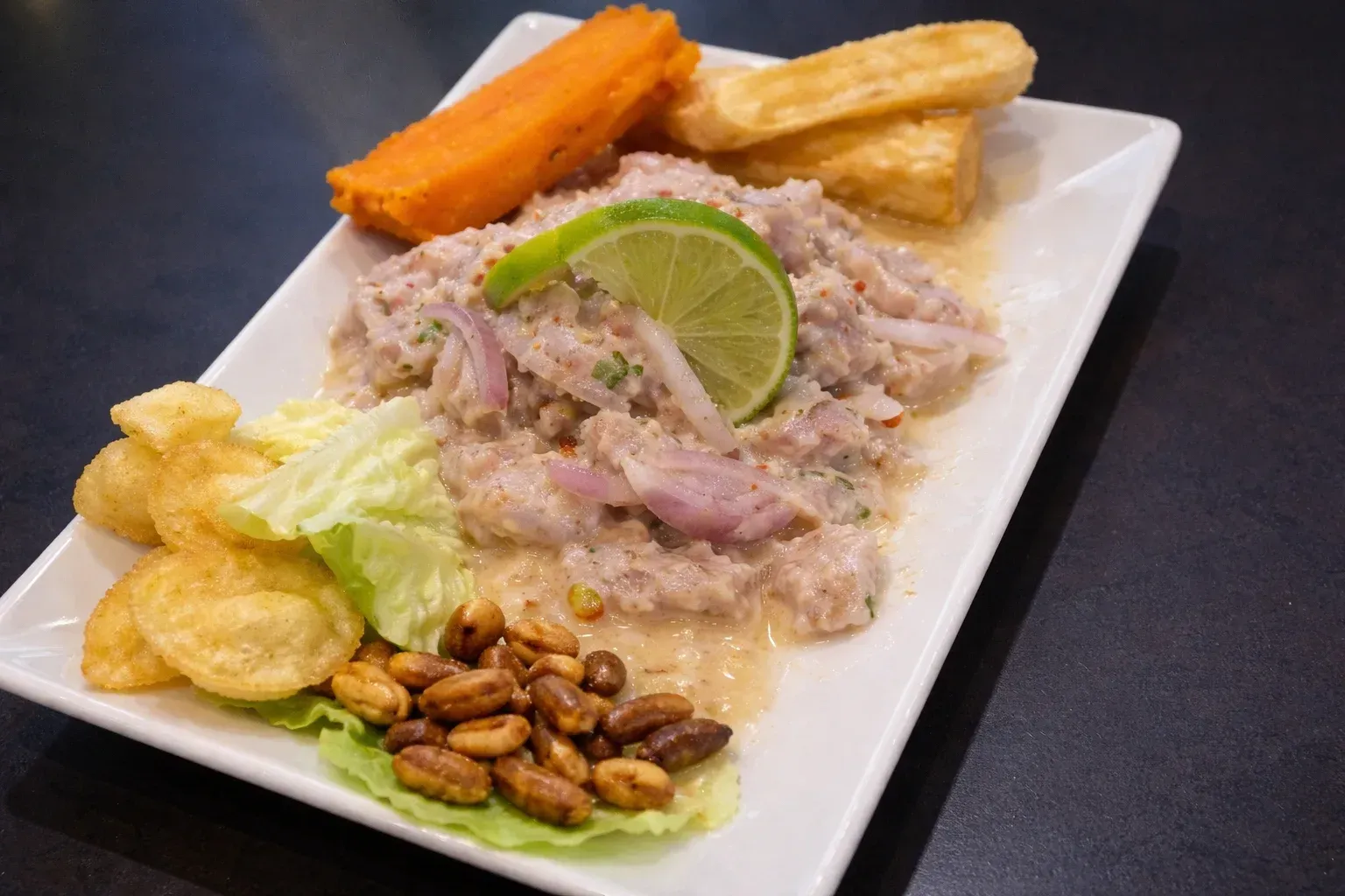 Plato de ceviche con limón, camote, yuca frita, maíz cancha y papas fritas en un plato blanco.