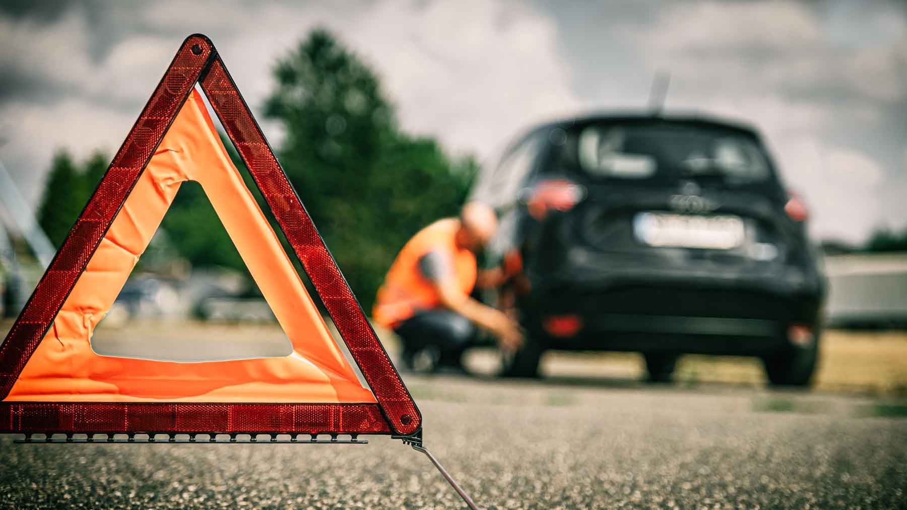 Un triangle de signalisation au premier plan avec un homme accroupi vérifiant le pneu arrière de son véhicule à l'arrêt