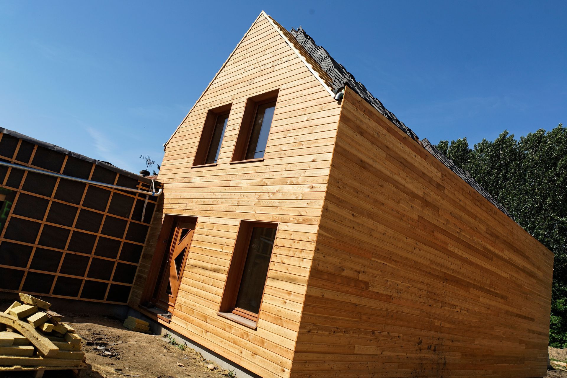 Maison en bois en construction avec un toit à pignon et des fenêtres, à l'extérieur par une journée ensoleillée.