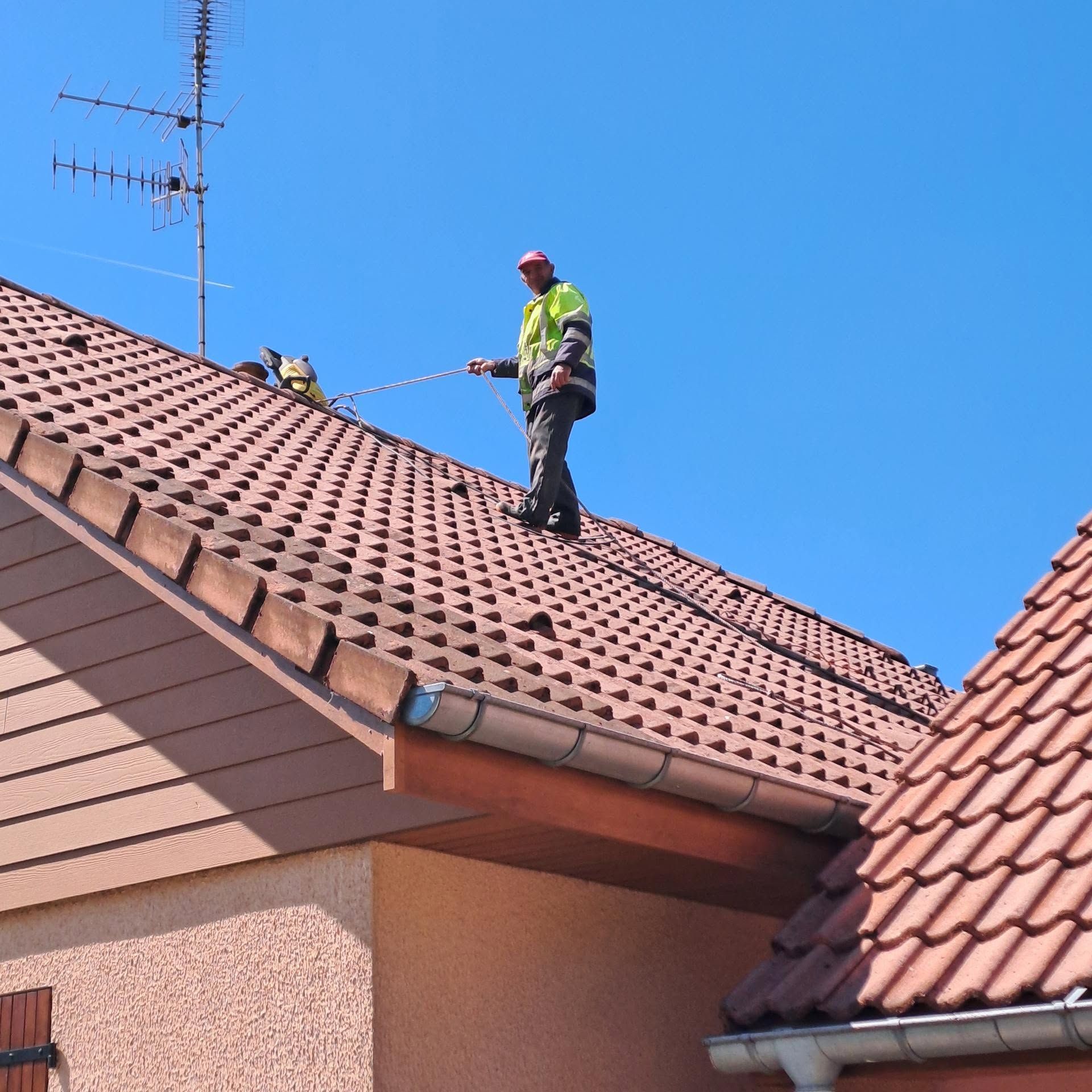 Un homme sur un toit en tuiles rouges nettoie ou inspecte les bardeaux sous un ciel bleu dégagé.