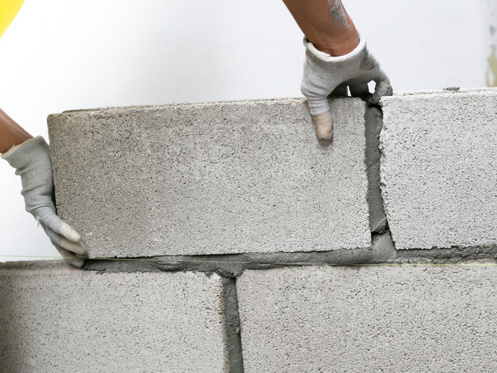Des mains avec des gants de travail gris posent un bloc de béton sur un mur en construction.