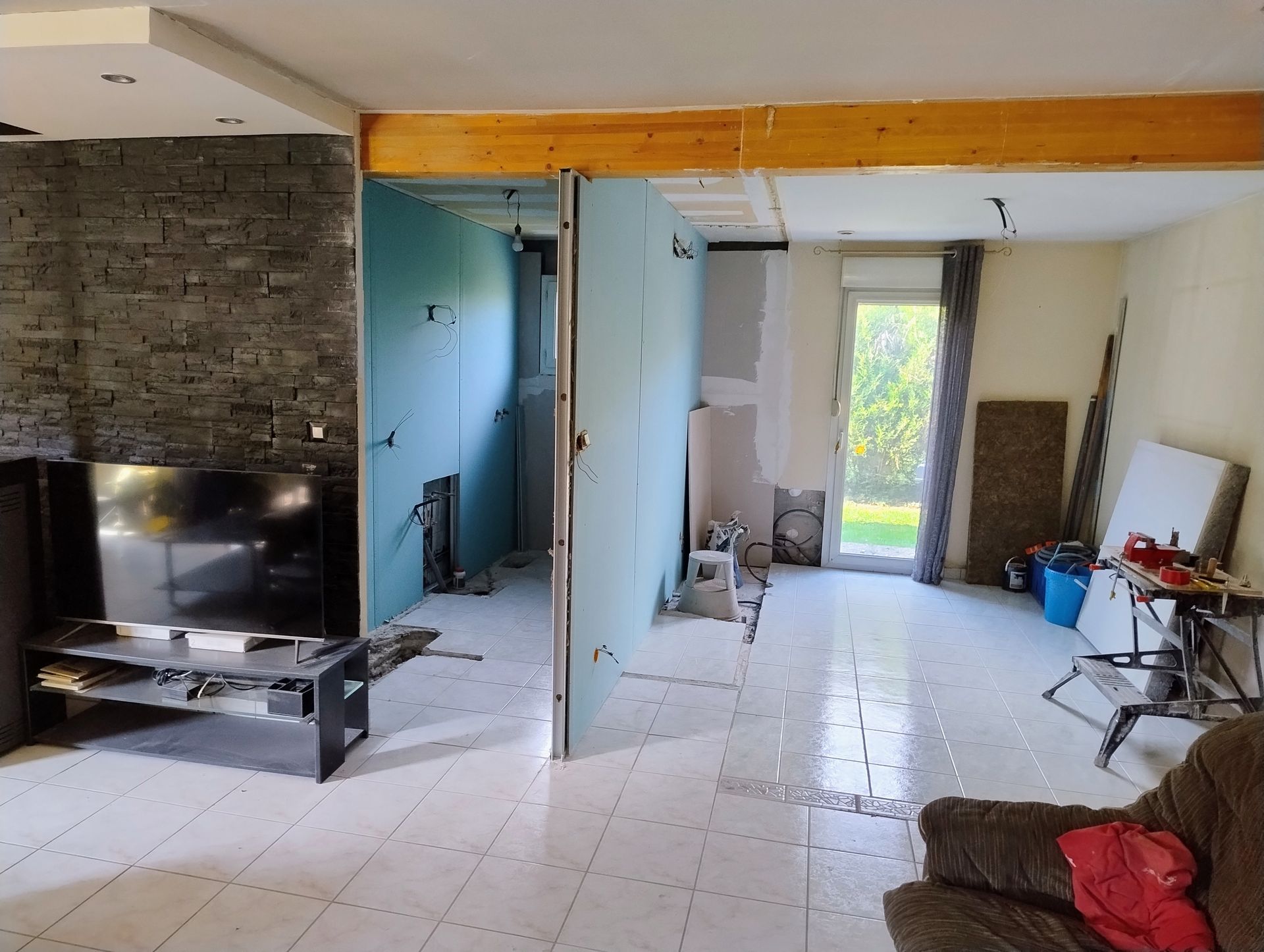 Salon rénové avec télévision, mur en pierre, sol carrelé, cloison et porte ouverte sur un patio lumineux.