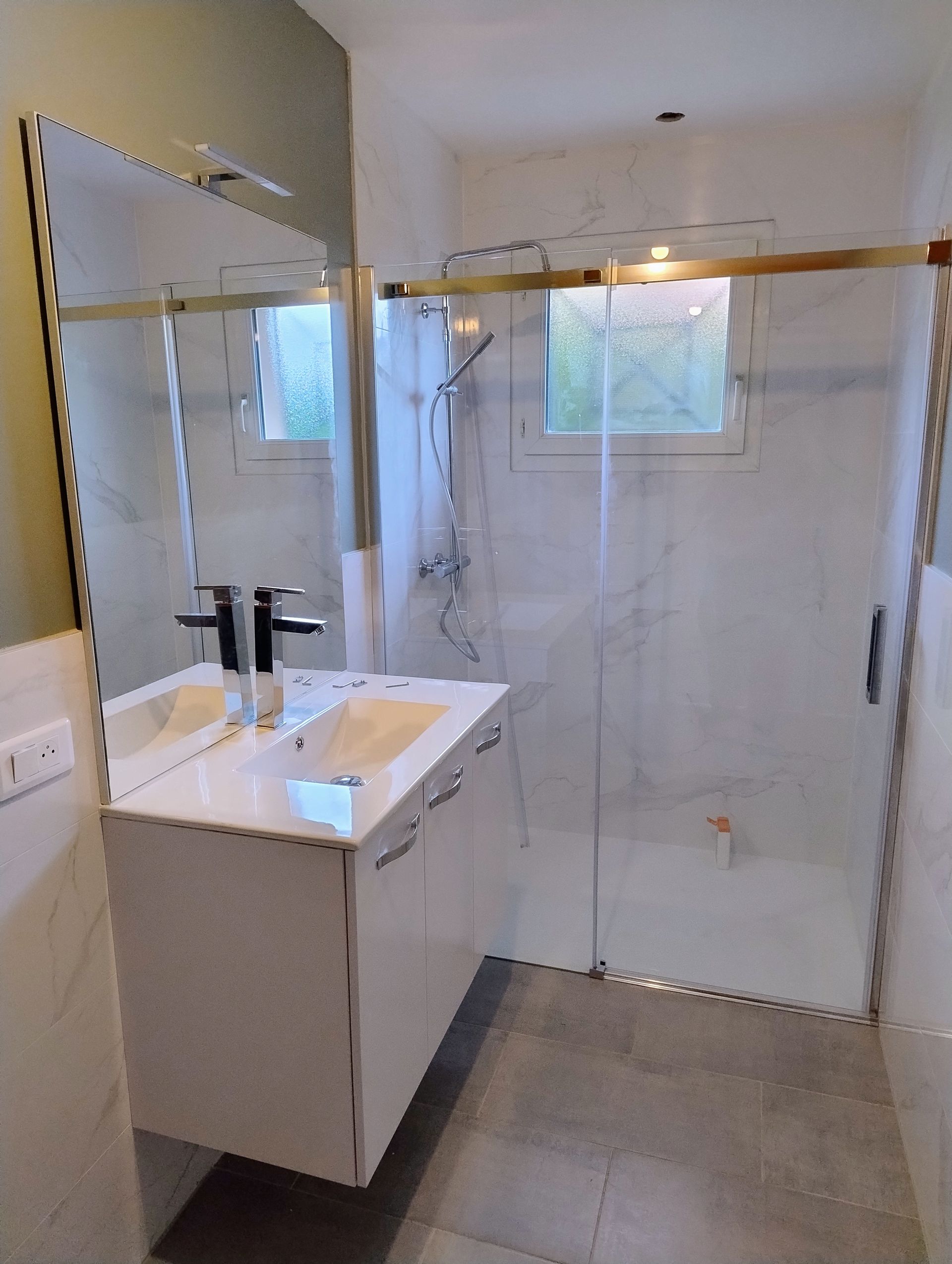 Salle de bains avec meuble-lavabo blanc, miroir, douche à l’italienne et sol carrelé gris.