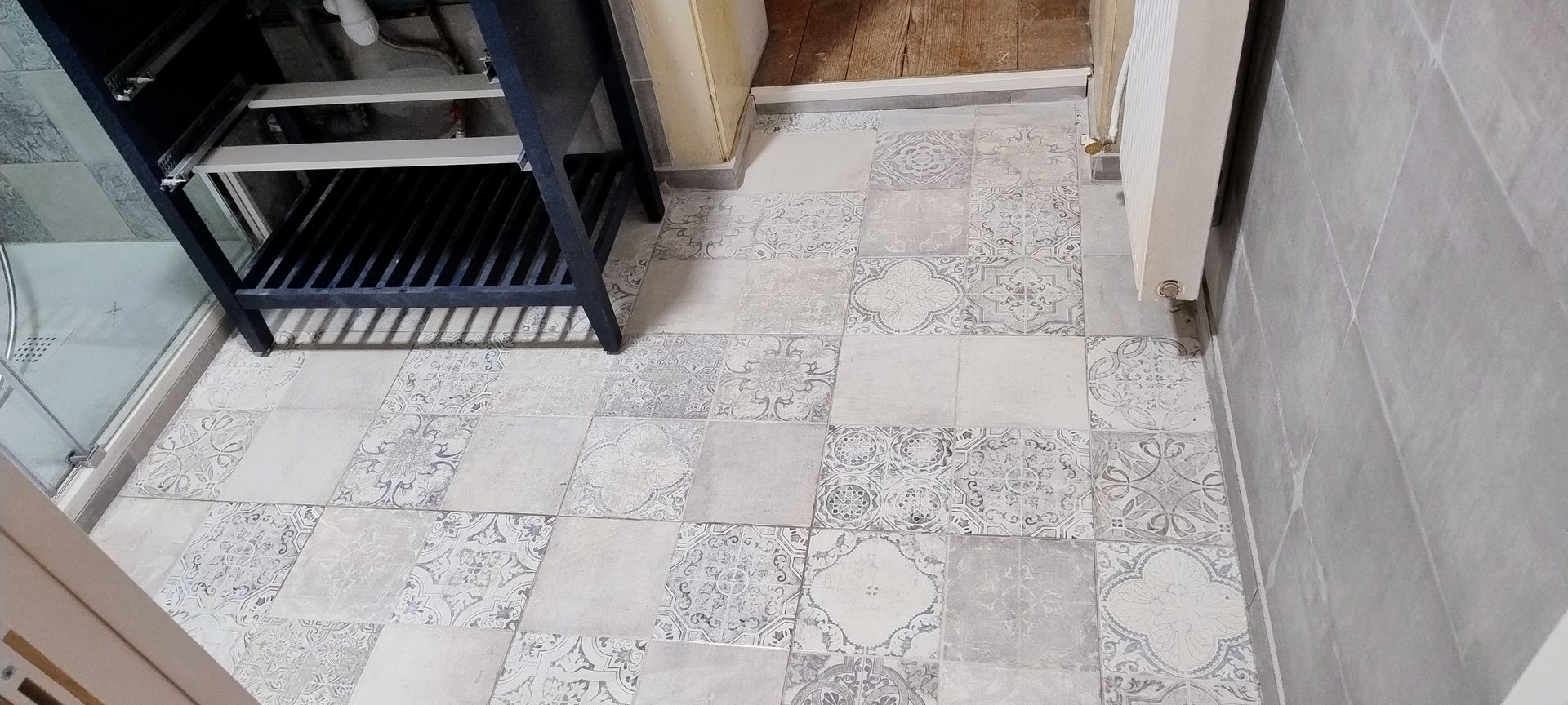 Petit sol de salle de bains carrelé, à côté d’une étagère noire et d’un mur beige.