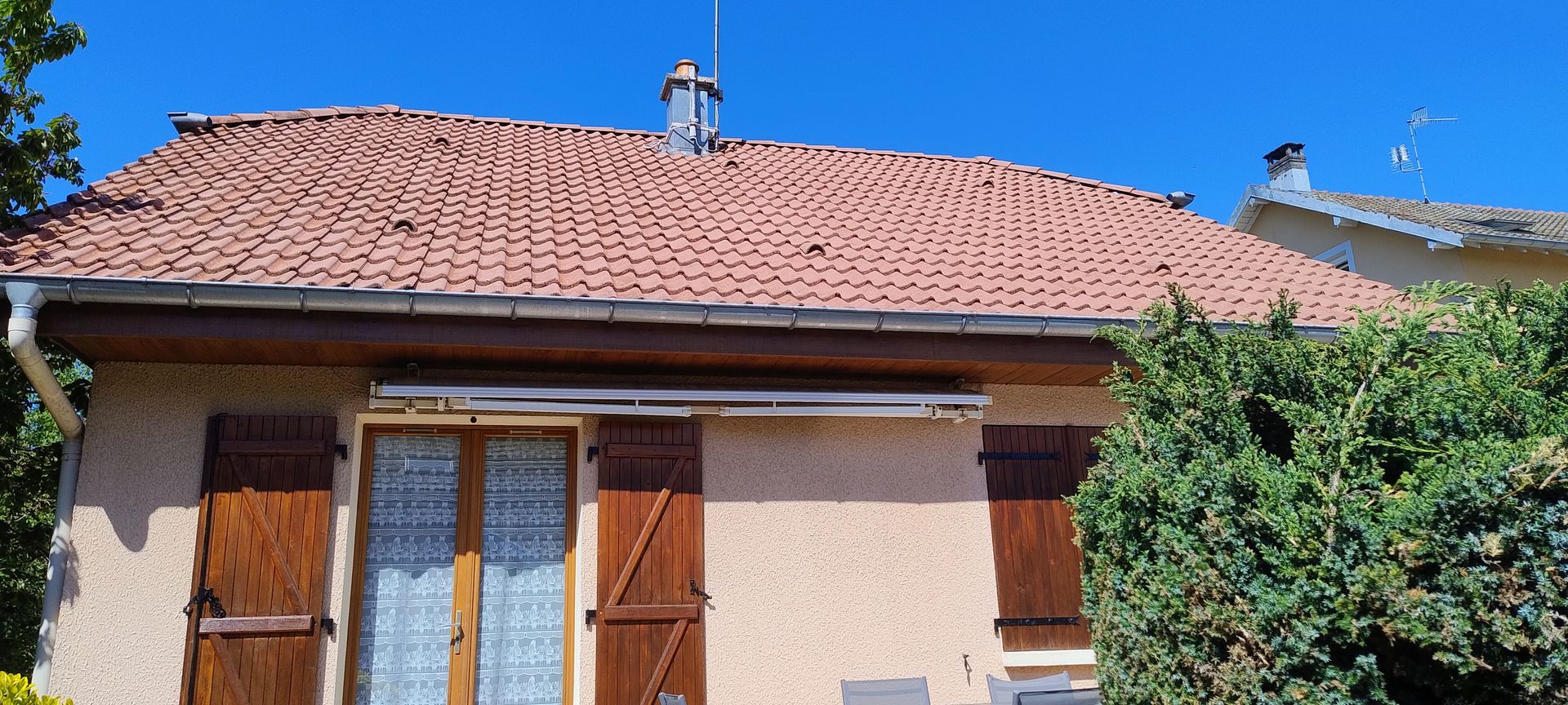Maison de plain-pied au toit de tuiles rouges, à la façade couleur pêche, avec des volets en bois et un arbuste feuillu à droite.