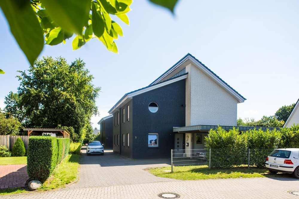 Modernes Haus mit dunkler und weißer Fassade, Hecken, geparkten Autos und einer hellen Auffahrt unter klarem Himmel