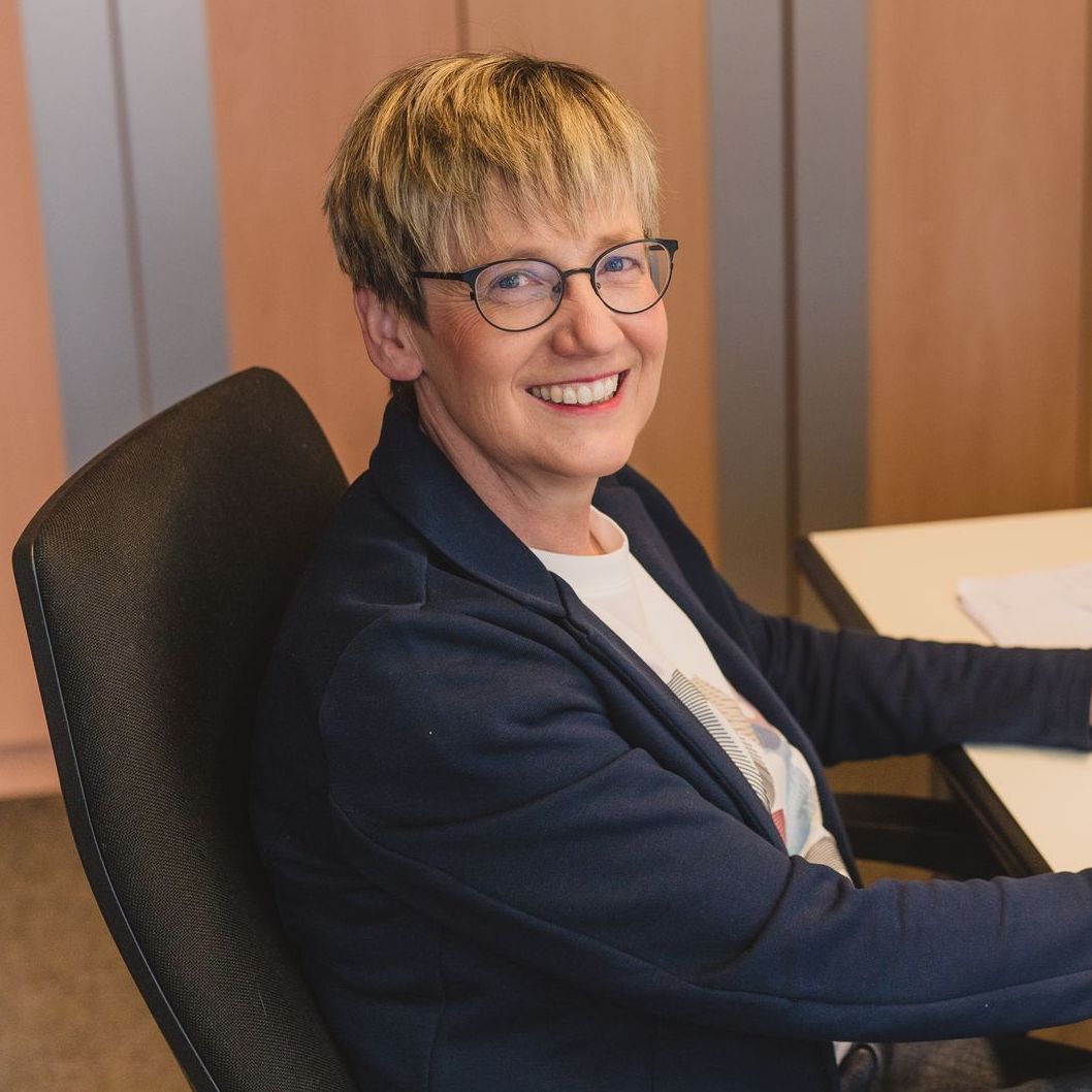 Eine Frau mit Brille lächelt, während sie an einem Schreibtisch sitzt