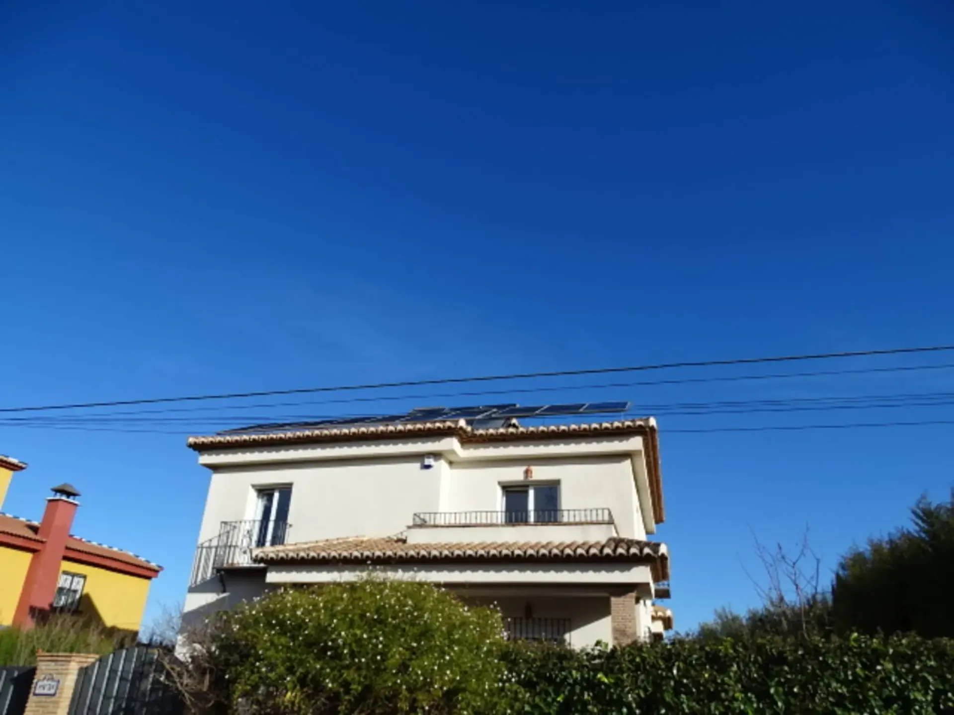 Casa de dos plantas de color crema con tejado de tejas, rodeada de arbustos bajo un cielo azul despejado.