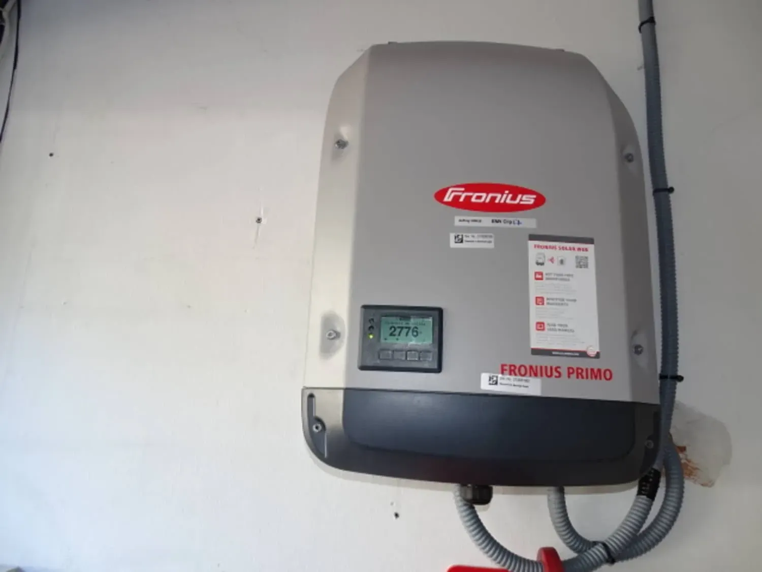 Inversor solar Fronius Primo gris montado en la pared con pantalla y cables sobre una pared blanca.