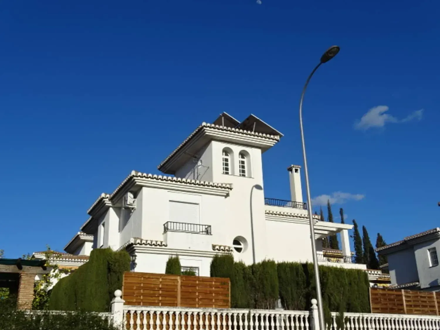 Villa blanca con tejado de tejas rojas y seto bajo un cielo azul despejado, vista desde la calle.