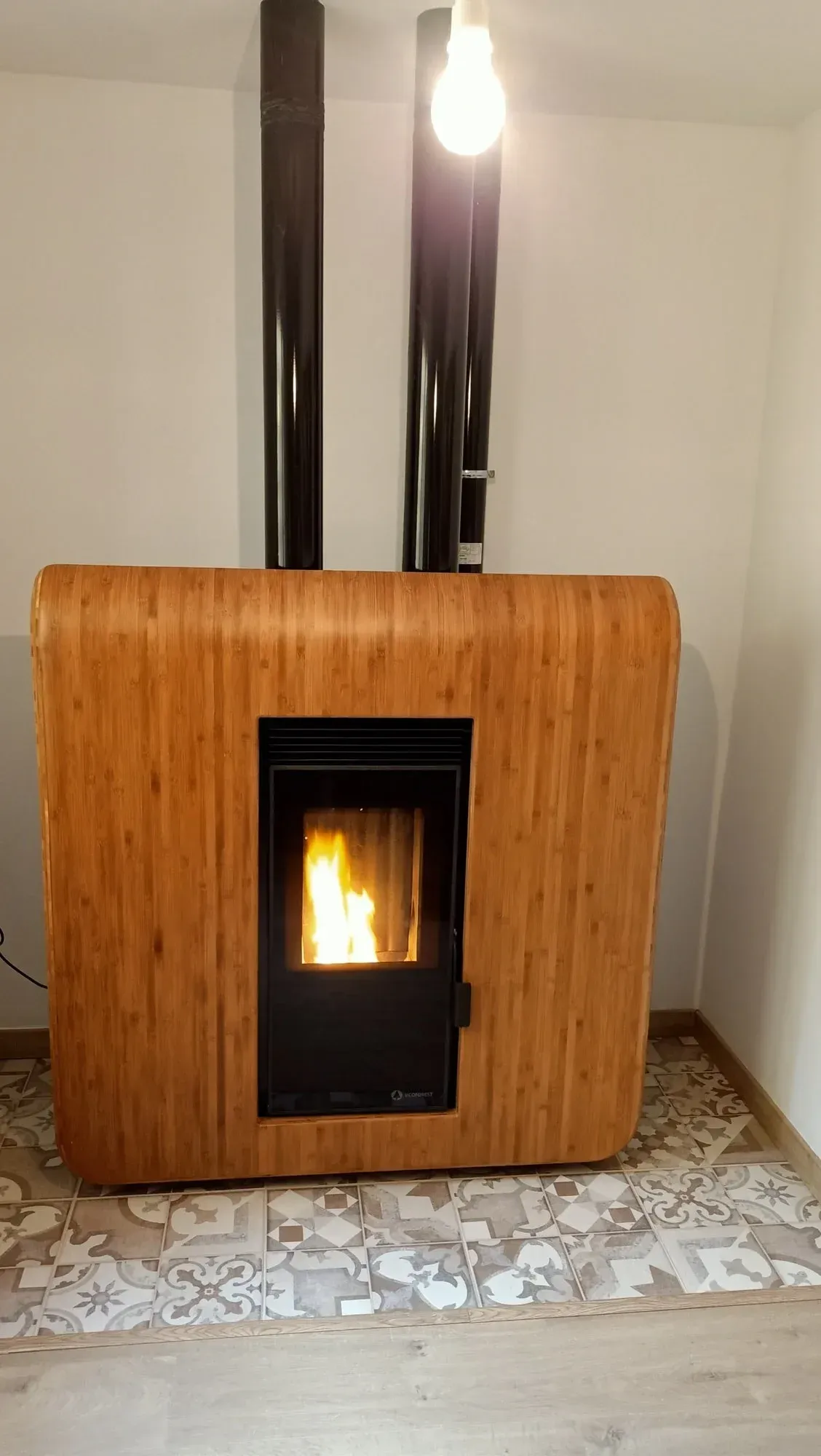 Estufa de leña con paneles de madera y fuego encendido, contra una pared clara sobre un suelo de baldosas estampadas.