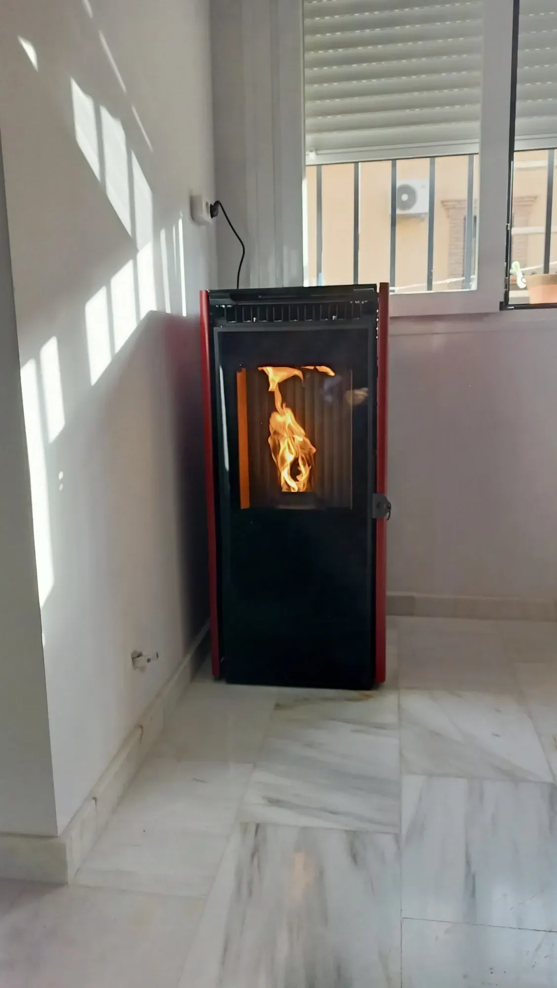 Estufa de leña negra con llama visible en una habitación luminosa con azulejos blancos cerca de una ventana.