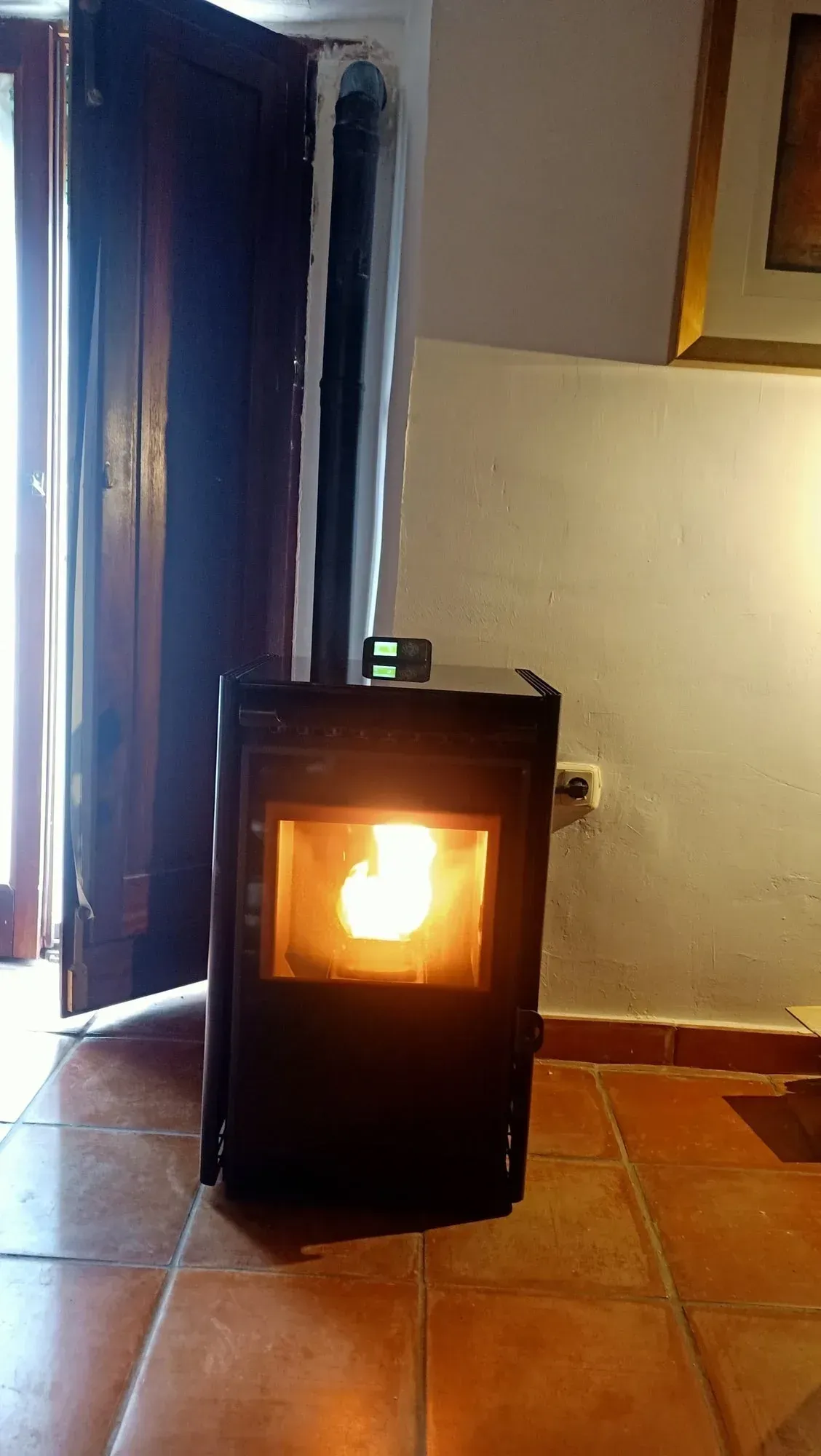 Una pequeña estufa de pellets negra, encendida, se encuentra en una habitación con azulejos cerca de una puerta.