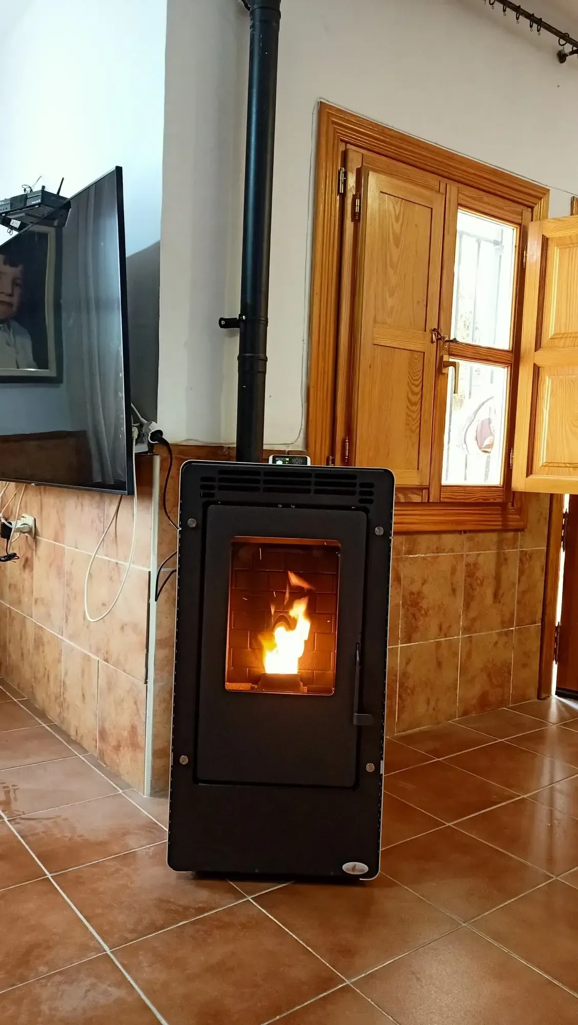 Estufa de leña negra con fuego encendido en una habitación con azulejos junto a una ventana y un televisor.