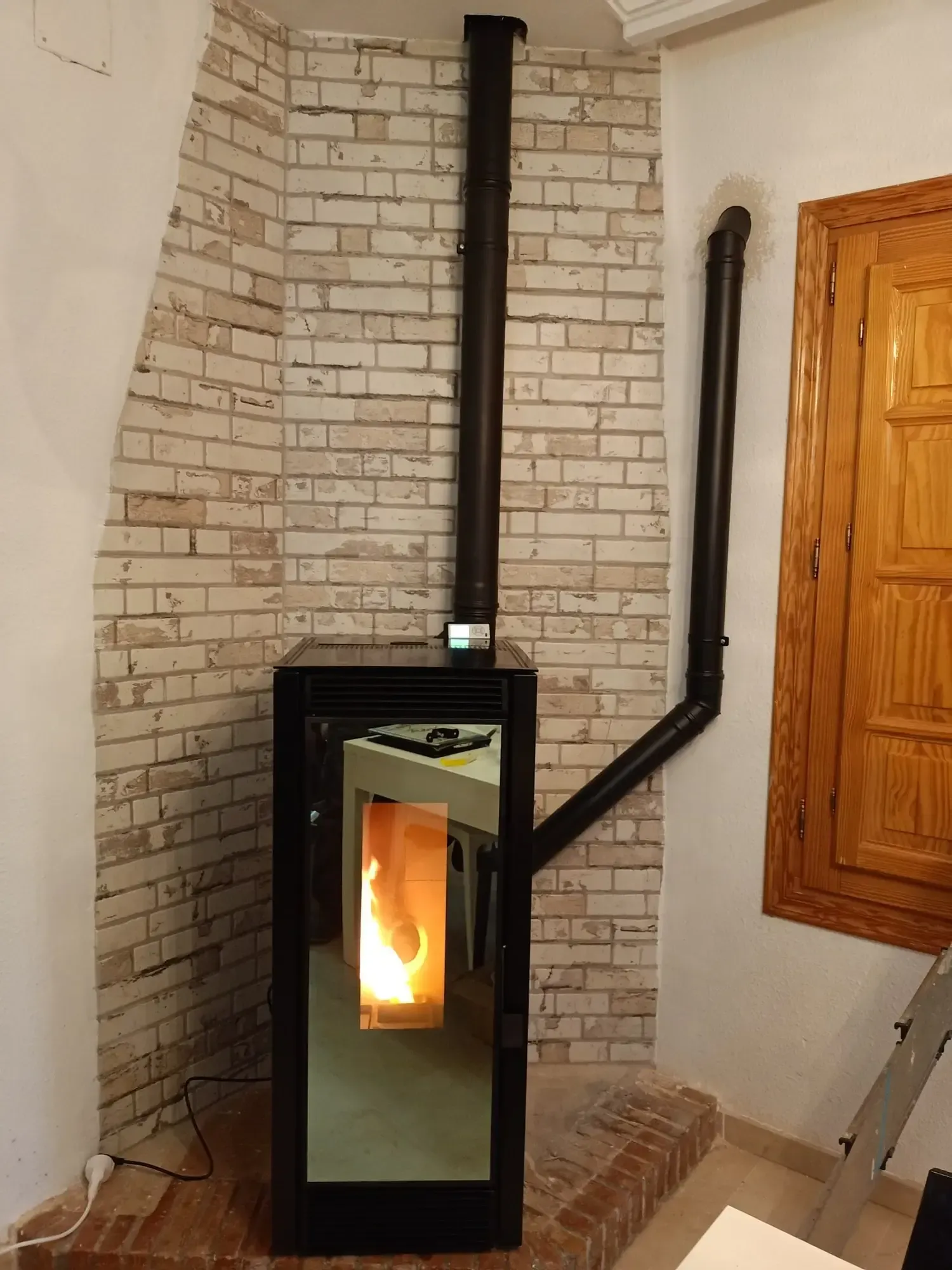 Estufa de leña negra con fuego brillante contra una pared curva de ladrillo blanco, con chimenea y frontal de espejo.