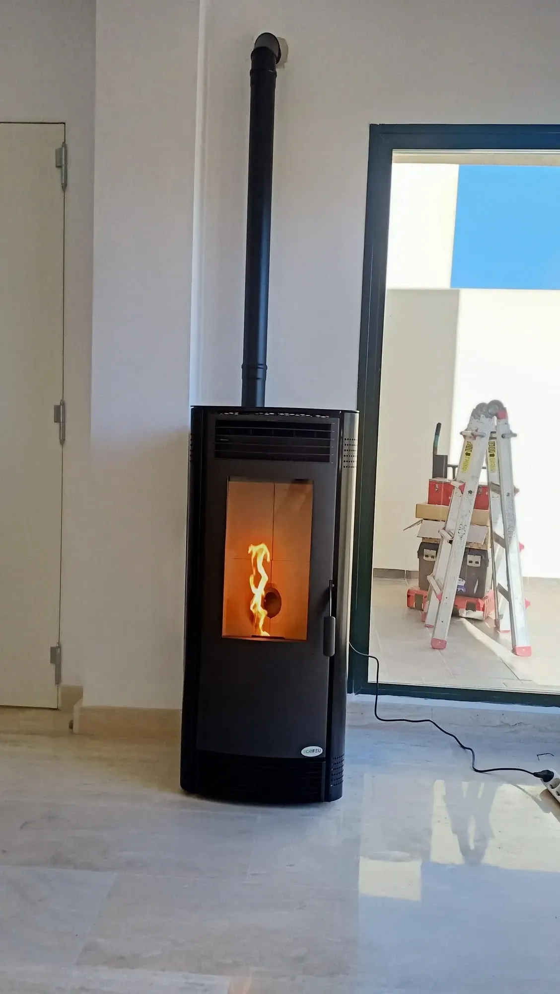 Estufa de leña negra con llama visible en una habitación luminosa, cerca de una puerta abierta y una escalera en el exterior.