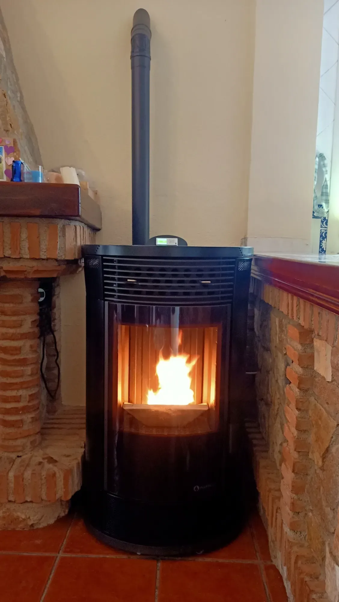 Estufa de pellets negra con fuego brillante en una habitación alicatada, centrada entre repisas de ladrillo.
