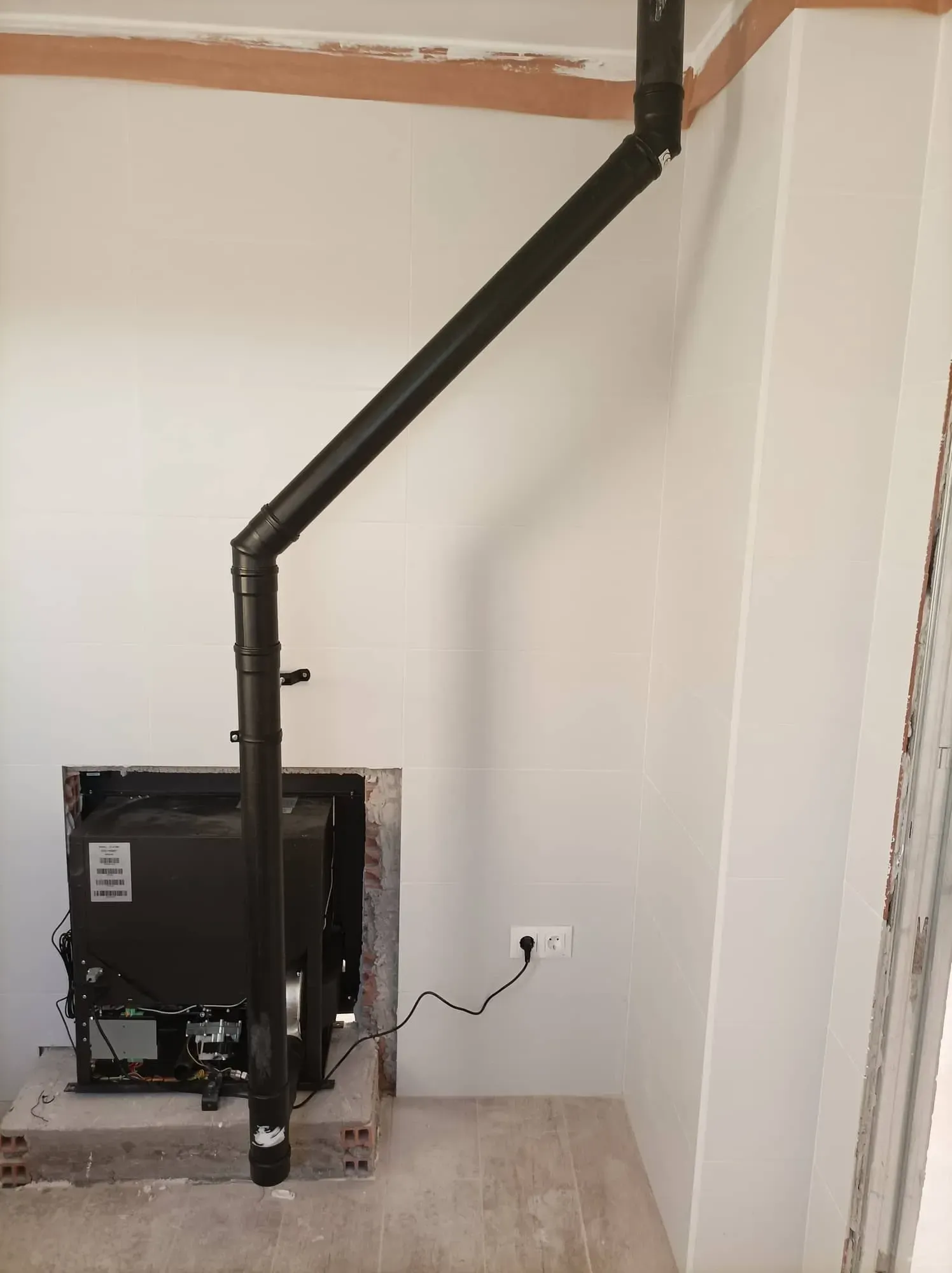 En una habitación blanca, una tubería de metal negro recorre diagonalmente desde una máquina en el suelo hasta la pared.