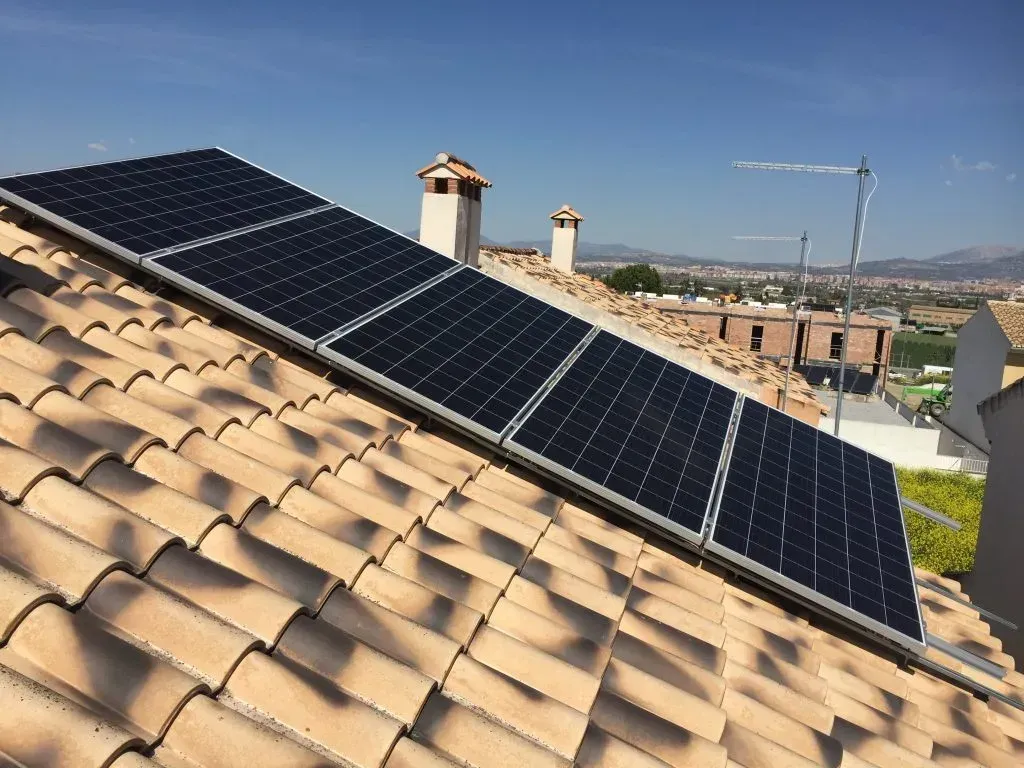Paneles solares en un tejado de tejas bajo un cielo azul despejado, con vistas a un barrio residencial.