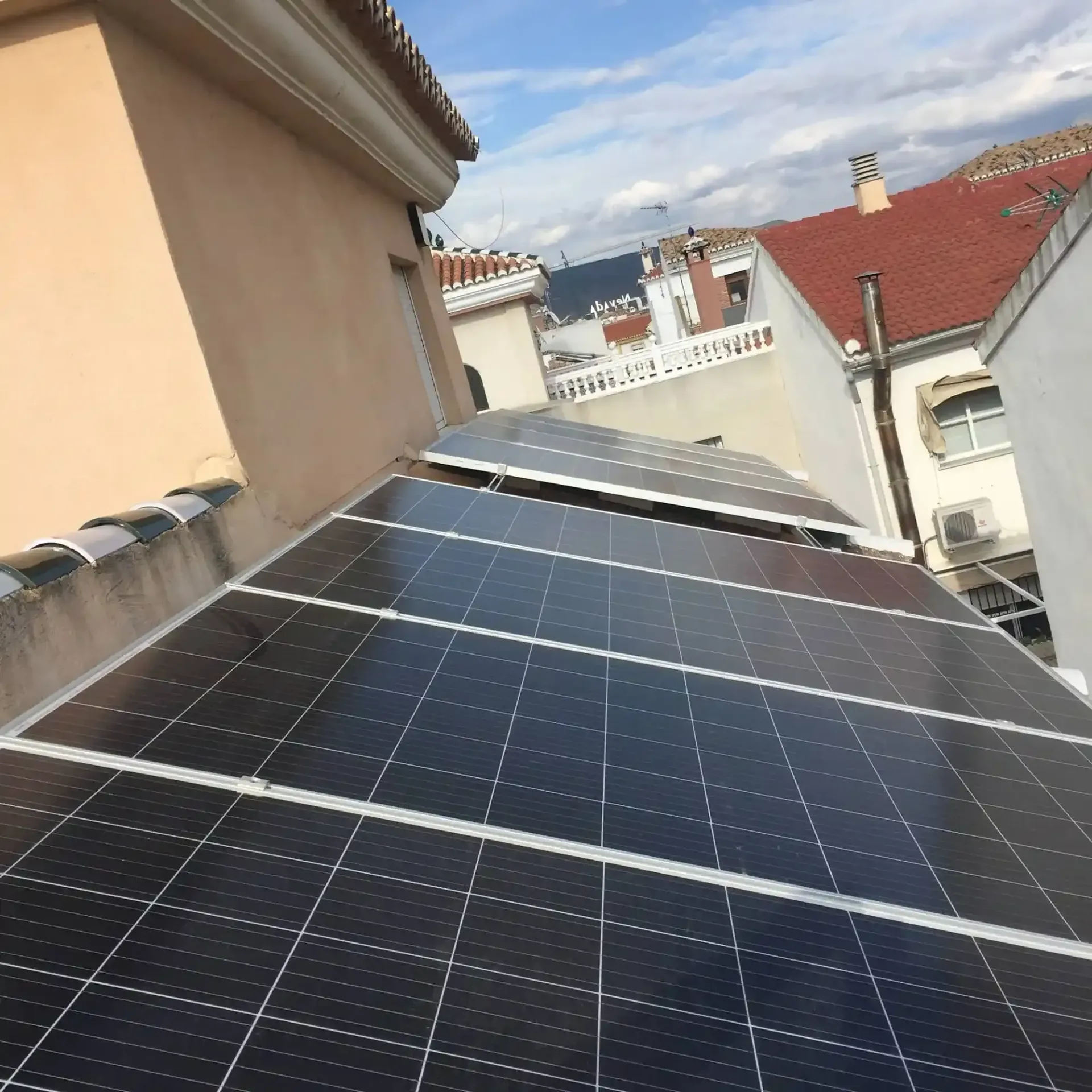 Paneles solares instalados en un tejado inclinado junto a edificios de tejados beige y rojos bajo un cielo nublado.