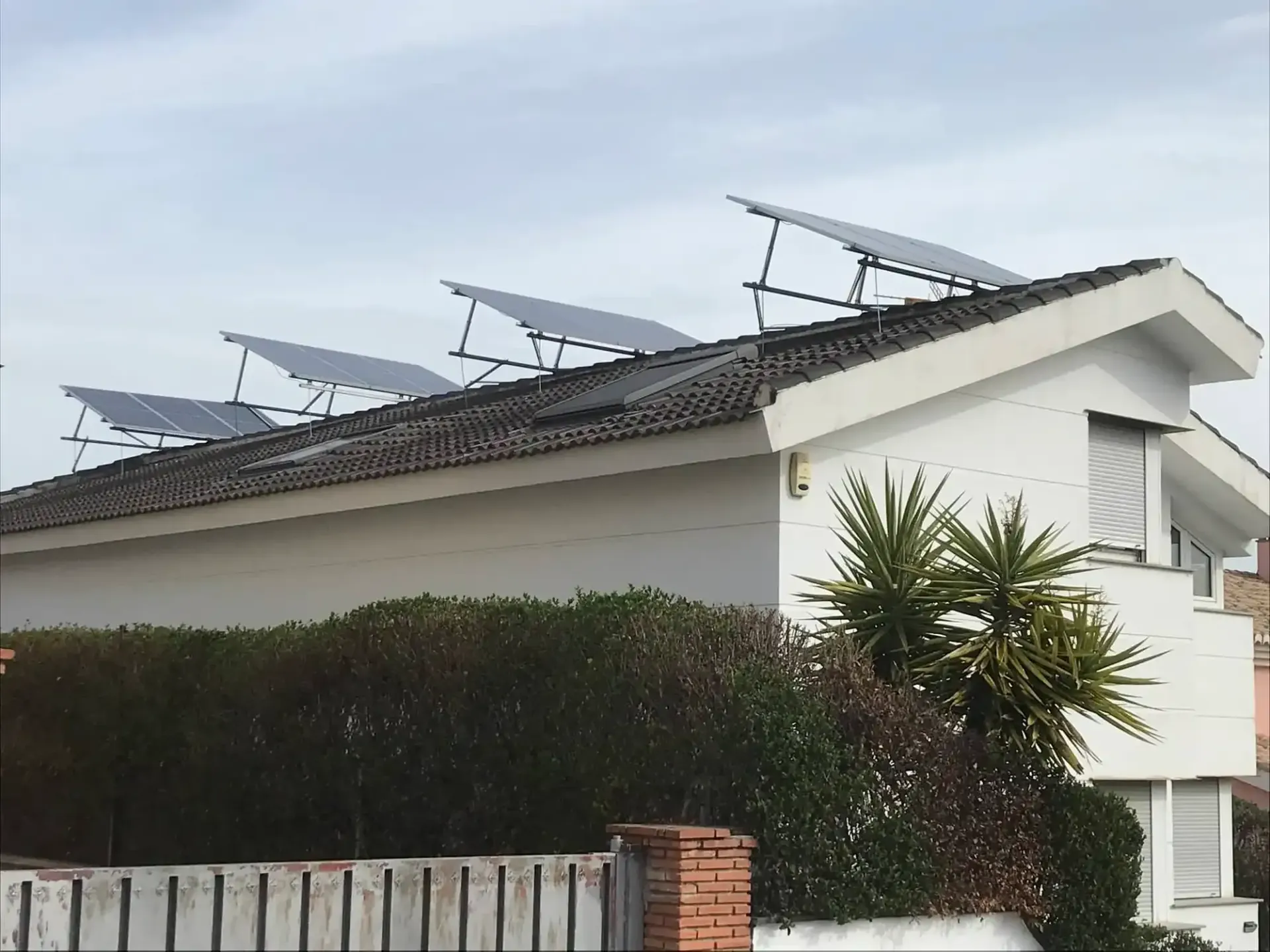 Techo blanco con paneles solares y antenas parabólicas detrás de un seto y una pared de ladrillos.